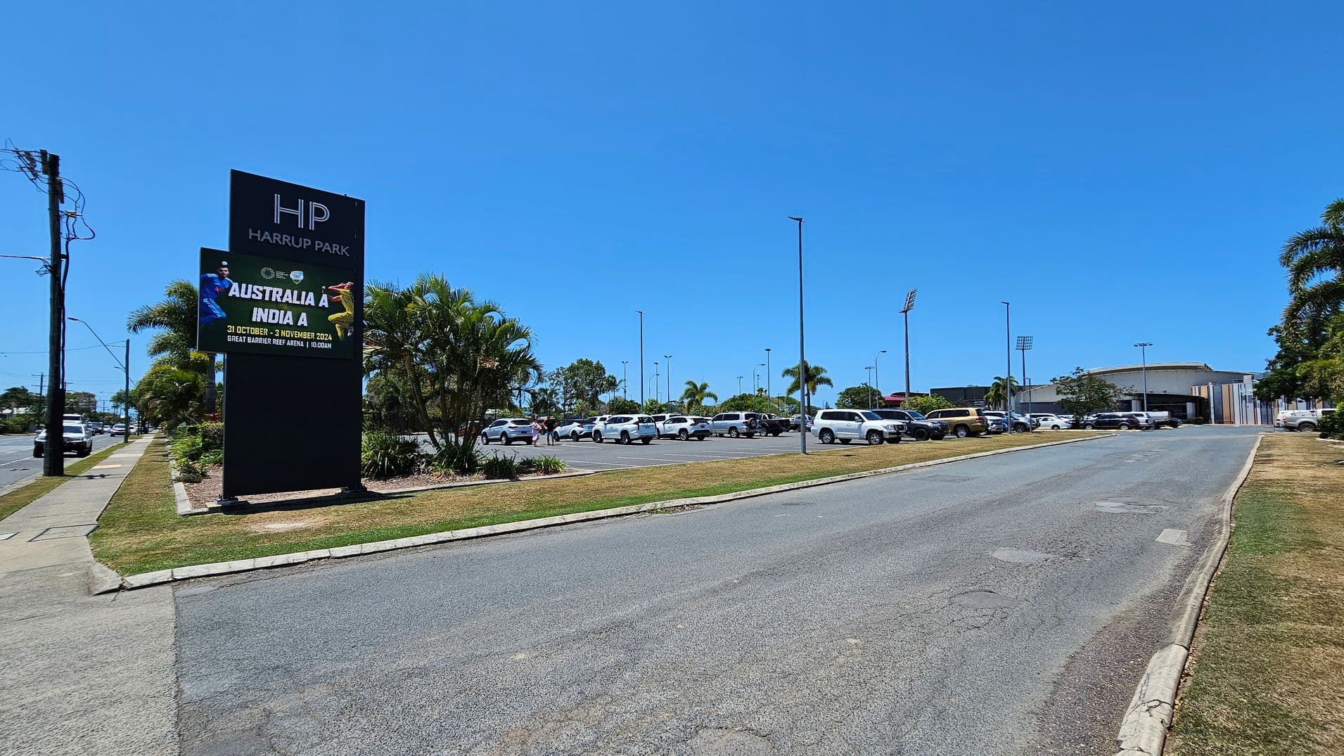 A road leads towards a parking area filled with cars on the right. On the left, a large sign reads "HP Harrup Park" with an advertisement for a cricket match between Australia A and India A. The sign is surrounded by palm trees and greenery. In the background, tall floodlights and a building are visible, likely part of a sports complex. The pavement is bordered by grass on both sides.