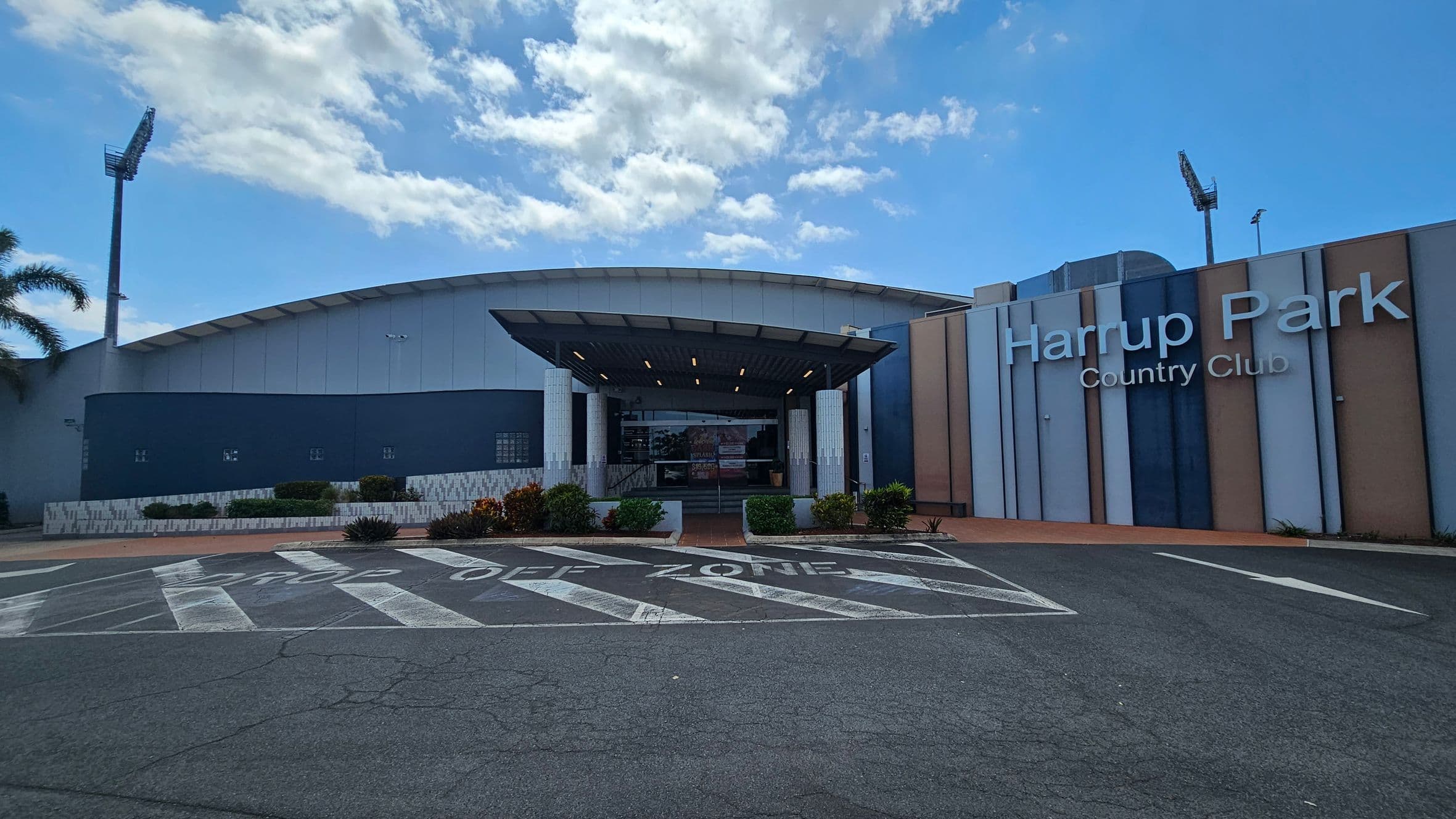 A modern building with a curved roof and a large entrance canopy. The facade features a mix of blue, grey, and beige panels. "Harrup Park Country Club" is prominently displayed on the right side. In front, there is a marked "Drop Off Zone" on the asphalt. The left side has a small garden with shrubs and a tall floodlight in the background.