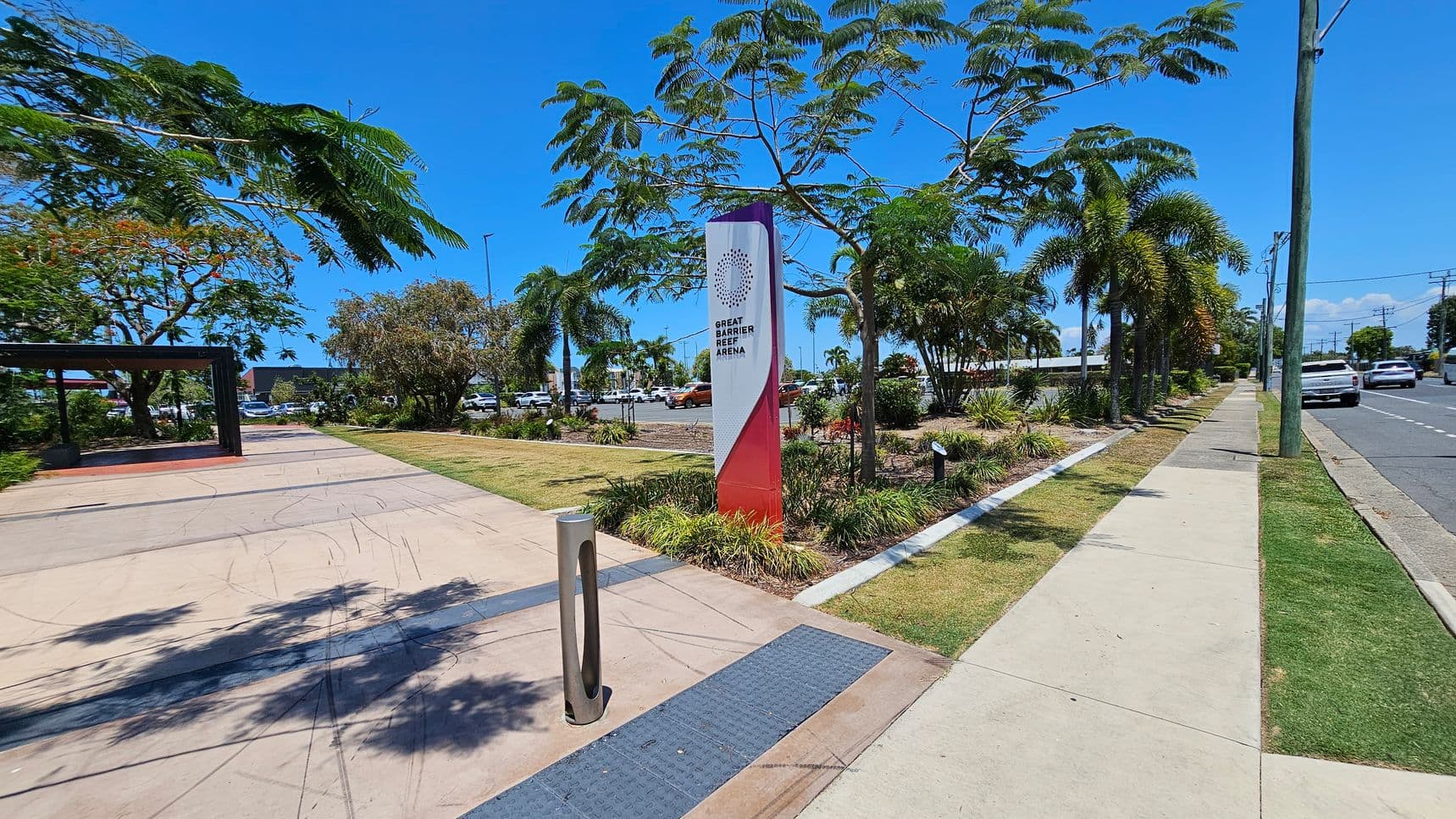 A paved walkway on the left with a metal bollard and a sign reading "Great Barrier Reef Arena" surrounded by greenery. Trees with lush foliage line the path. On the right, a road with cars and a footpath bordered by grass and palm trees. A utility pole stands near the road. The area is well-maintained with a mix of concrete and grassy surfaces.