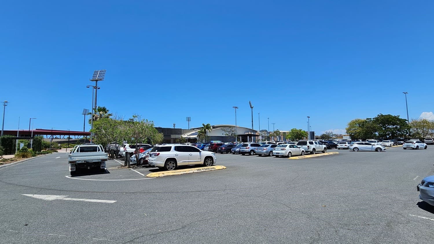 A large car park with numerous vehicles, including white and silver cars, is in the foreground. In the background, there is a stadium with tall floodlights and a curved roof. Trees and small shrubs are scattered around the parking area. A red pedestrian bridge is visible on the left side, and the ground is paved with asphalt.