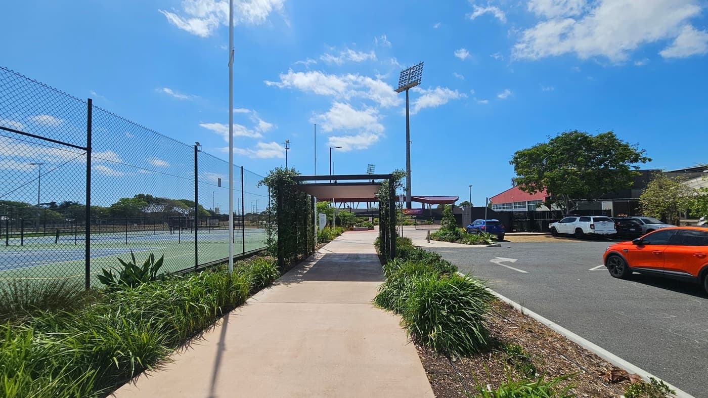 A pathway runs through the centre, bordered by green plants and leading to a structure with a roof. On the left, a chain-link fence encloses tennis courts. On the right, a car park with several vehicles, including a prominent orange car, is visible. A large tree provides shade near the parked cars. Tall floodlights are visible in the background, and the sky is clear with a few clouds.