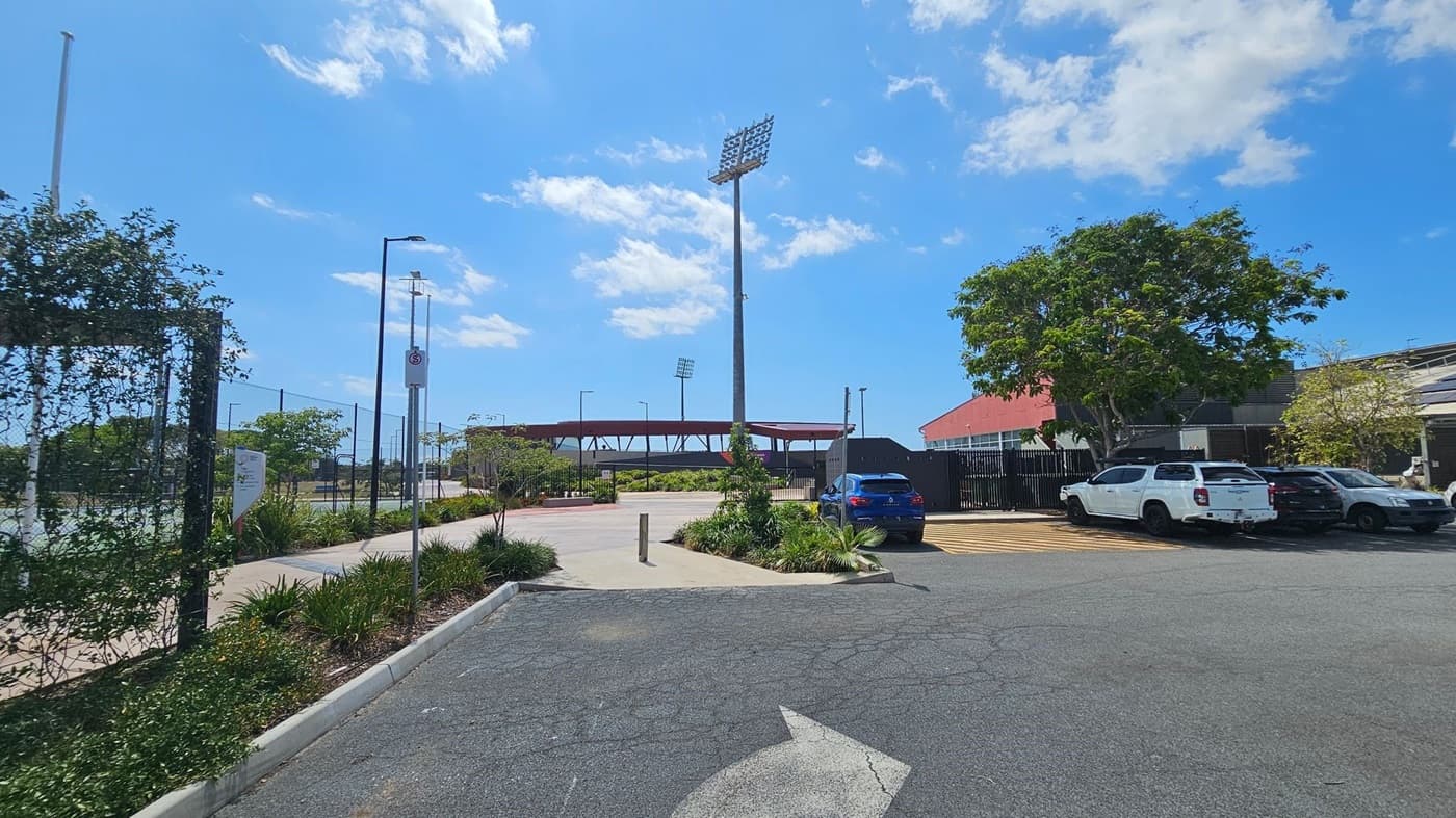 A car park with several vehicles on the right, including a blue car and a few white ones. A large tree provides some shade. In the centre, a tall floodlight tower stands near a sports field, with a red-roofed structure in the background. On the left, a fenced area with greenery and a pathway runs alongside it. The ground is paved with visible markings, and a directional arrow points forward.