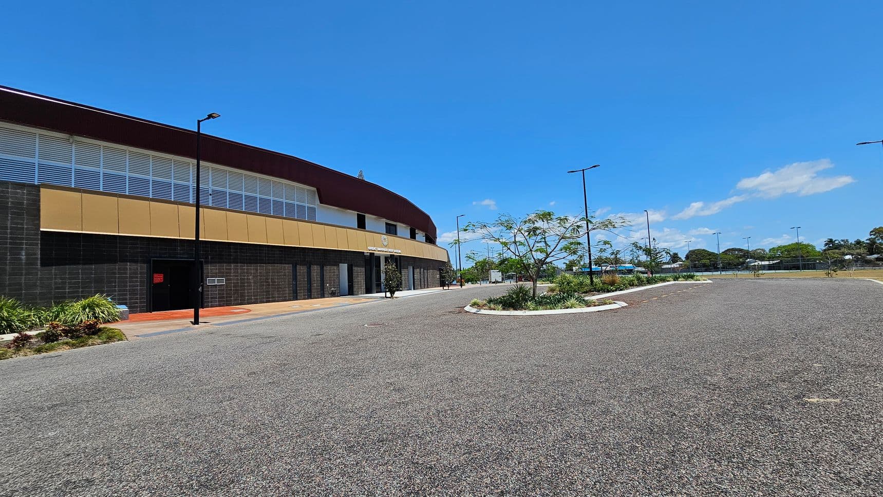 A large, modern building with a curved roof on the left, featuring a combination of dark grey and beige panels. The building has a row of windows near the top. In front, there's a paved walkway with a few small plants. On the right, a wide, empty asphalt road curves gently, bordered by landscaped areas with young trees and shrubs. Several tall streetlights line the road, and in the background, there are more trees and a fenced area.