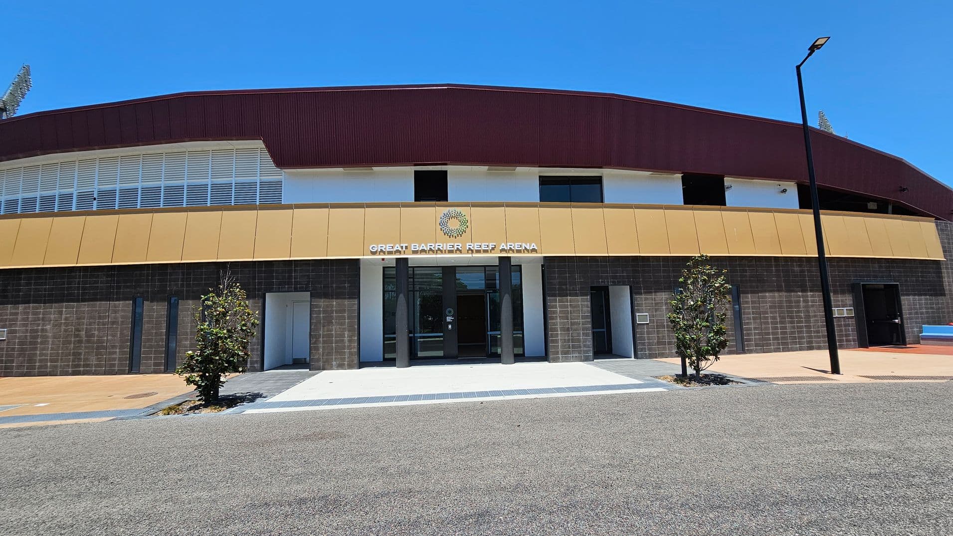 A modern building with a brown roof and a beige facade, featuring the sign "Great Barrier Reef Arena" above the entrance. The lower part of the building is clad in dark grey tiles. Two small trees are planted in front, flanking the entrance. A tall streetlamp stands to the right. The ground is paved with a mix of grey and beige surfaces.