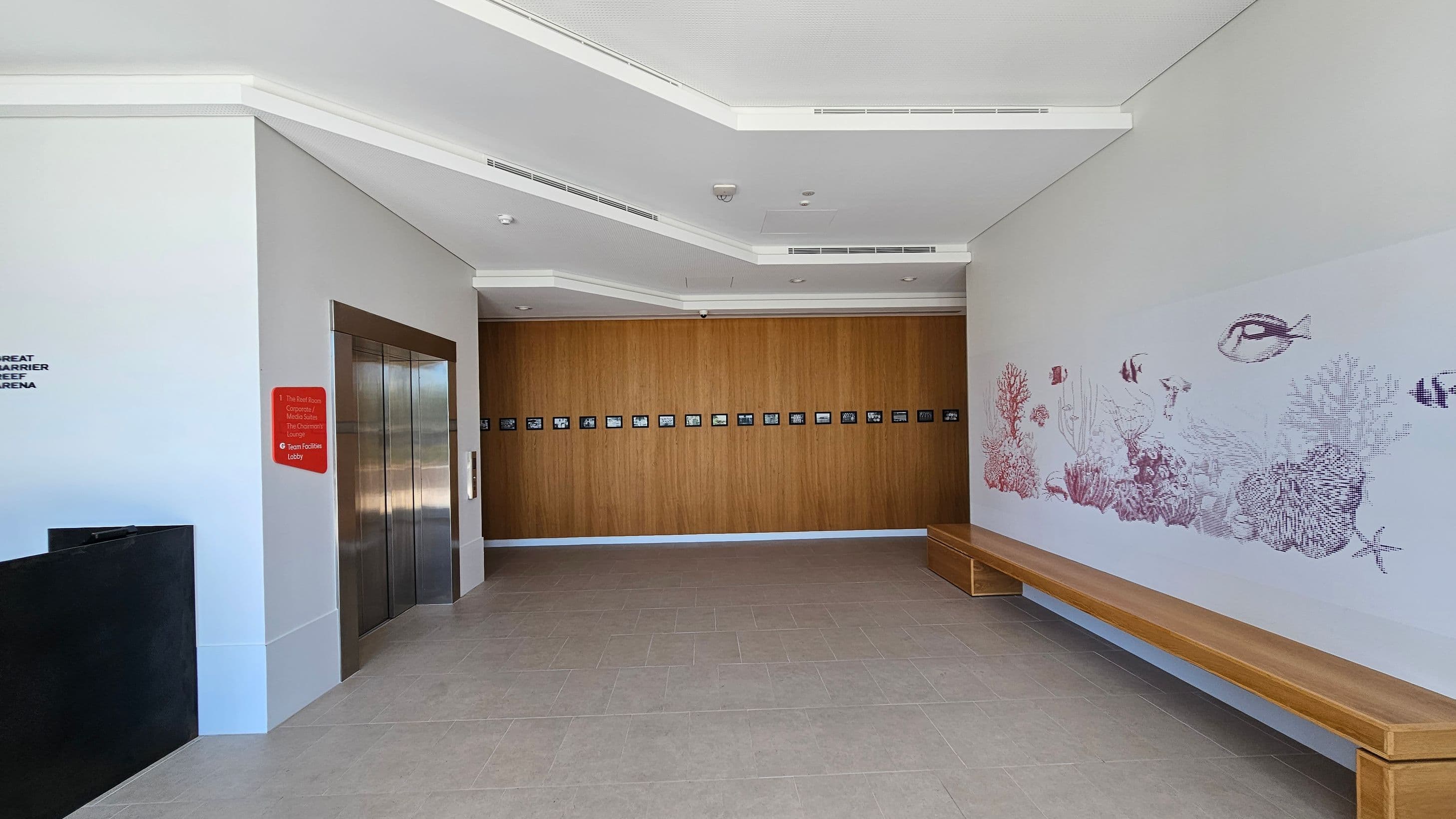 A modern interior space with a light-coloured tiled floor and a white ceiling featuring recessed lighting. On the left, there are two metallic lift doors with a red sign listing destinations. The back wall is wooden with a row of small framed pictures. On the right, a long wooden bench is positioned against a wall adorned with a detailed mural of marine life, including fish and coral.