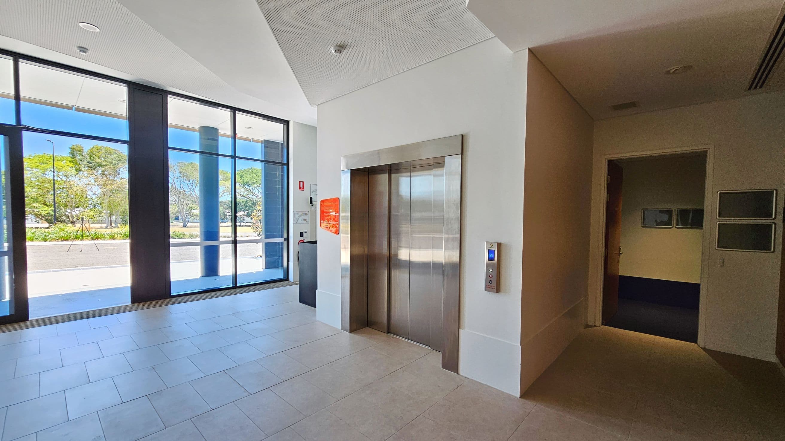On the left, large glass windows reveal a view of trees and a pathway outside. In the middle, a metallic lift with closed doors is set into a white wall. To the right, a dimly lit hallway leads to a room with framed pictures on the wall. The floor is tiled in a light colour, and the ceiling is white with recessed lighting.