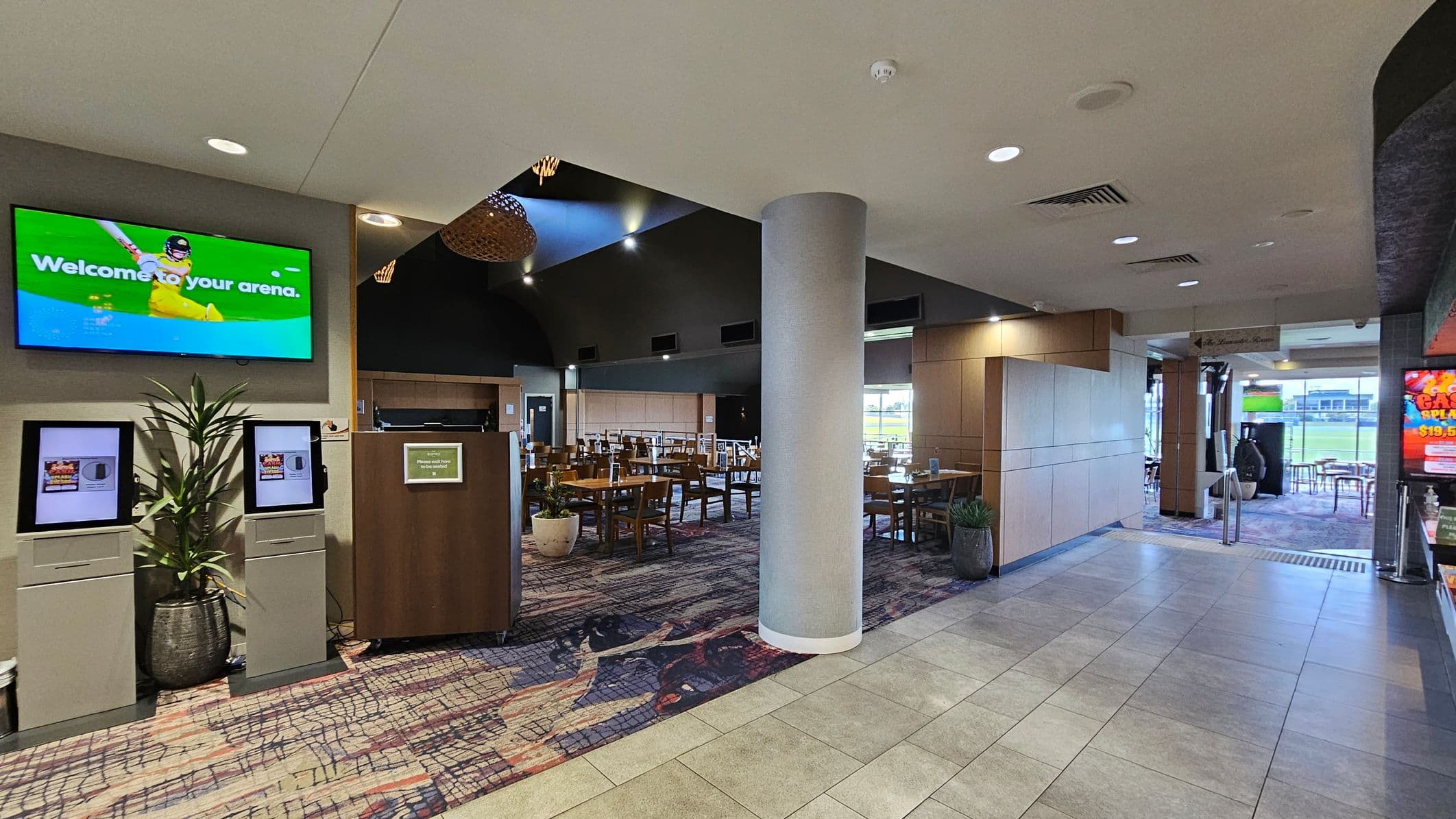 A spacious indoor area with a modern design. On the left, a large screen displays a welcome message above two kiosks and a potted plant. In the centre, a column stands in front of a dining area with wooden tables and chairs. The floor is a mix of patterned carpet and grey tiles. On the right, a bright digital sign is visible near the entrance, which leads to an outdoor area with more seating. The ceiling is fitted with recessed lighting and air vents.
