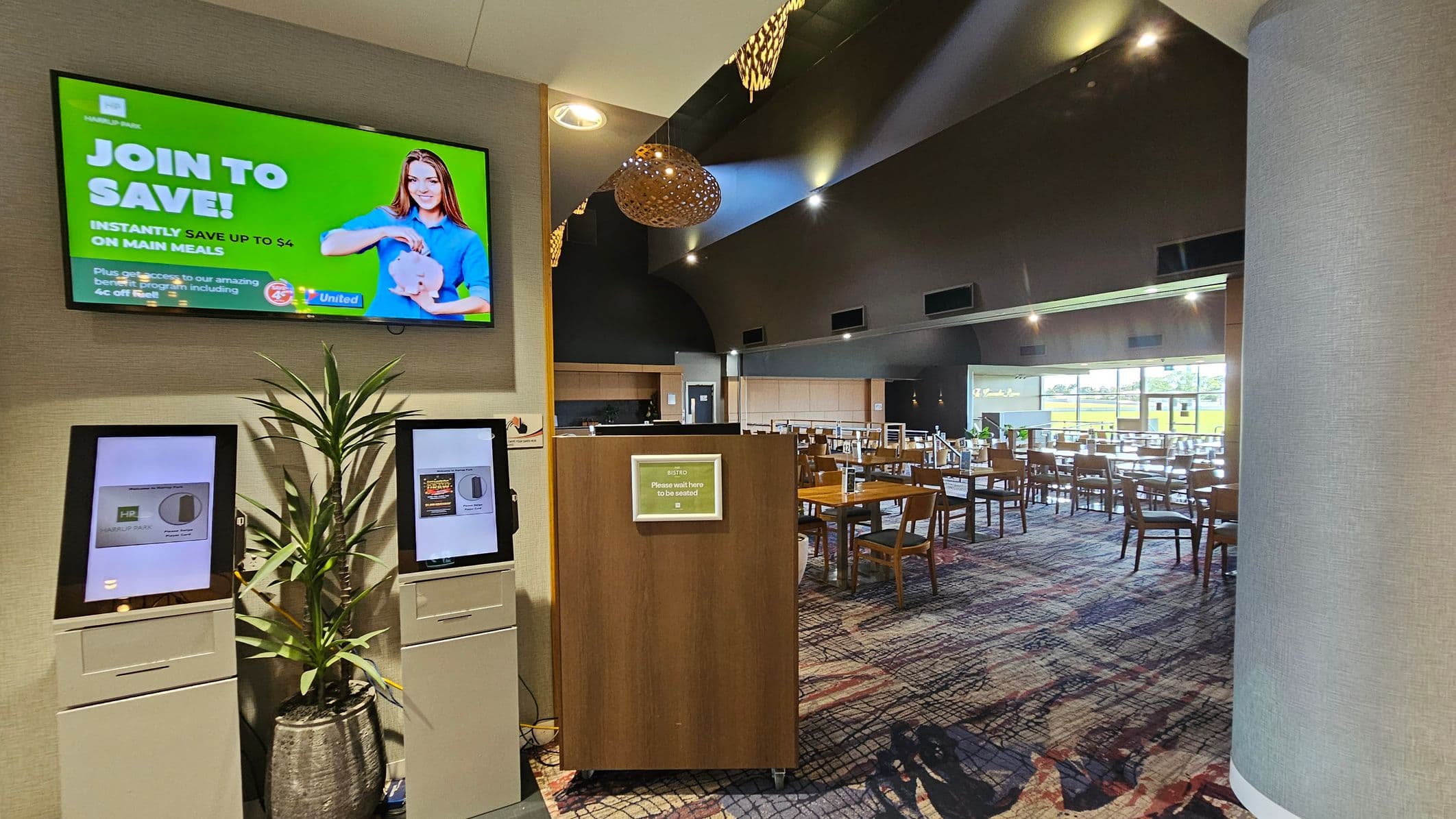 On the left, a digital screen displays a green advertisement with a person holding a piggy bank. Below it, two touchscreen kiosks stand beside a potted plant. In the middle, a wooden stand with a sign instructs visitors to wait. On the right, a spacious dining area with wooden tables and chairs is visible, with large windows in the background. The ceiling is high with modern lighting, and the floor is covered in a patterned carpet.