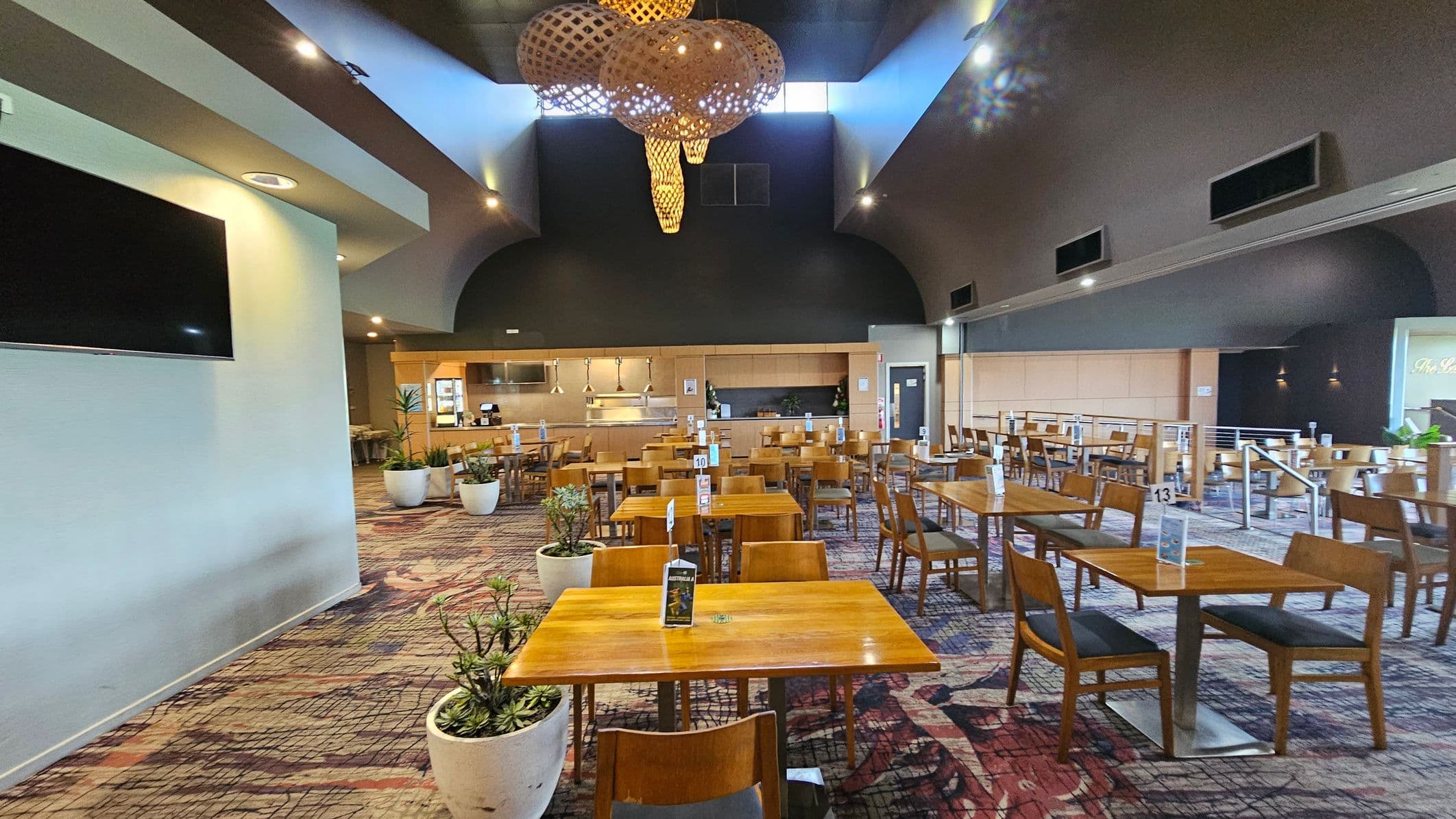 A spacious dining area with wooden tables and chairs arranged neatly. The floor is covered with a patterned carpet. Large potted plants are placed around the room. The ceiling is high with intricate wooden light fixtures. On the left, a large flat-screen TV is mounted on a light-coloured wall. The back wall features a serving counter with kitchen equipment visible. The right side has more seating and a partially visible sign. The lighting is bright and evenly distributed.