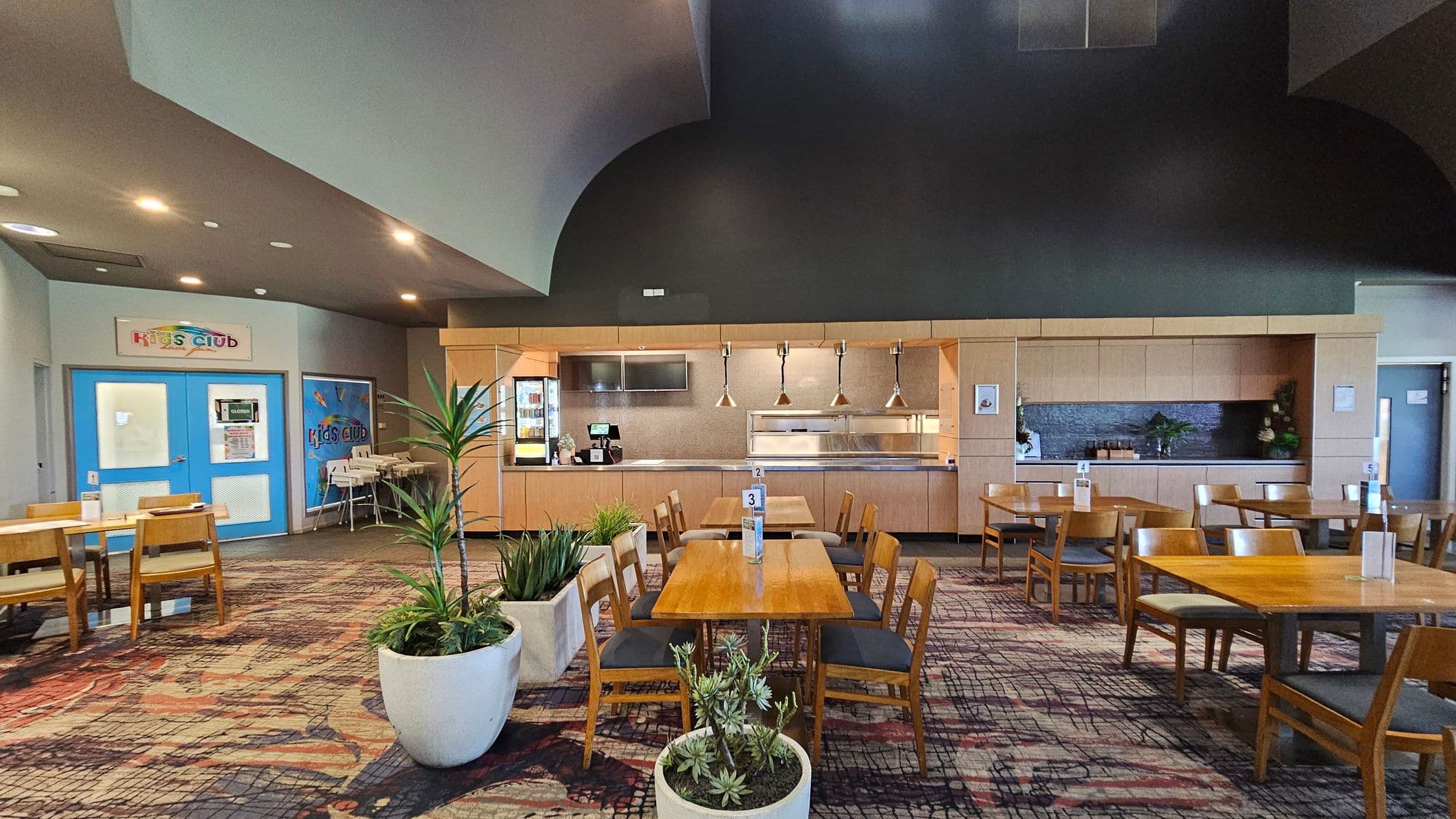 A spacious dining area with wooden tables and chairs arranged neatly. In the foreground, there are large potted plants. The background features a counter with hanging lights and a service area. On the left, a blue door marked "Kids Club" is visible, with high chairs nearby. The carpet has a patterned design, and the ceiling is high with recessed lighting.