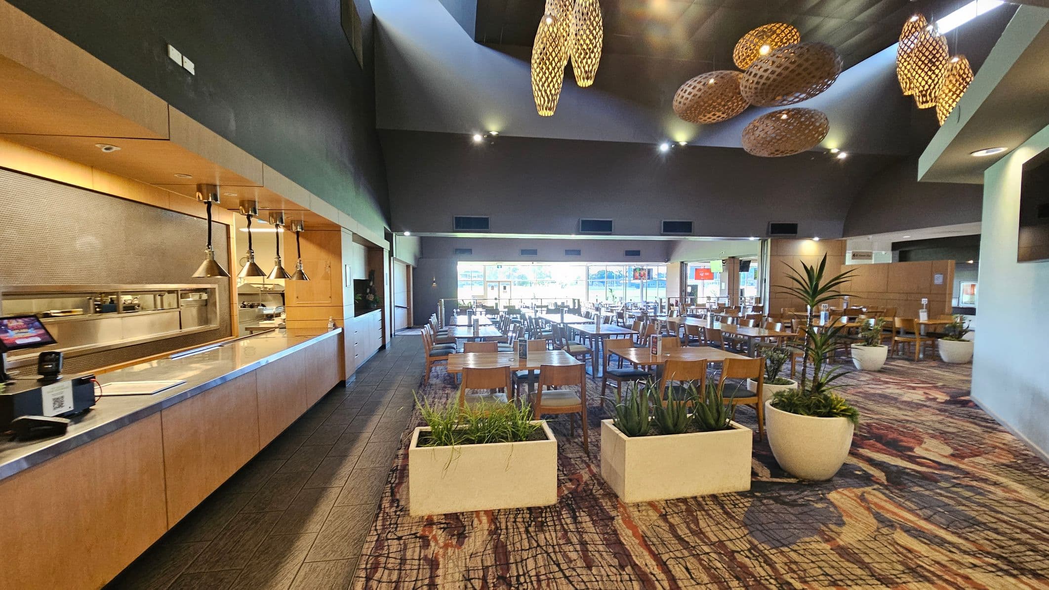 A spacious dining area with wooden tables and chairs arranged neatly. On the left, there's a serving counter with hanging lamps and a cash register. The floor is covered with a patterned carpet, and large potted plants are placed throughout. The ceiling is high with decorative woven light fixtures. At the back, large windows allow natural light to fill the room, and the walls are painted in neutral tones.
