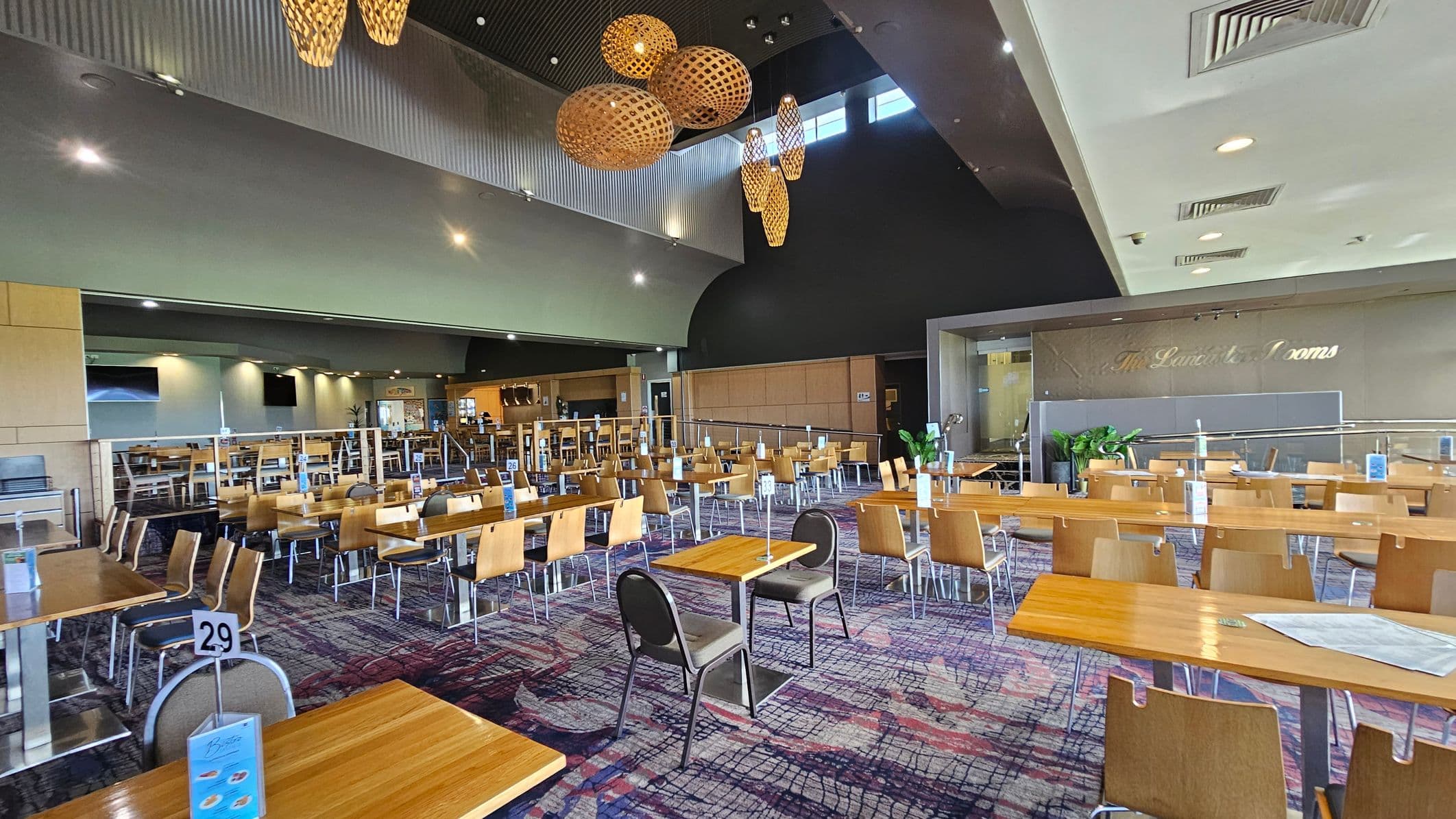 A spacious dining area with numerous wooden tables and chairs arranged in rows. The floor is covered with a patterned carpet in shades of blue and red. On the left, there are more tables and a menu stand with the number 29. The ceiling is high with large, woven light fixtures hanging down. In the background, a sign reads "The Lunch Room" on a wall, and there are potted plants near the entrance. The ceiling has recessed lighting and air vents.