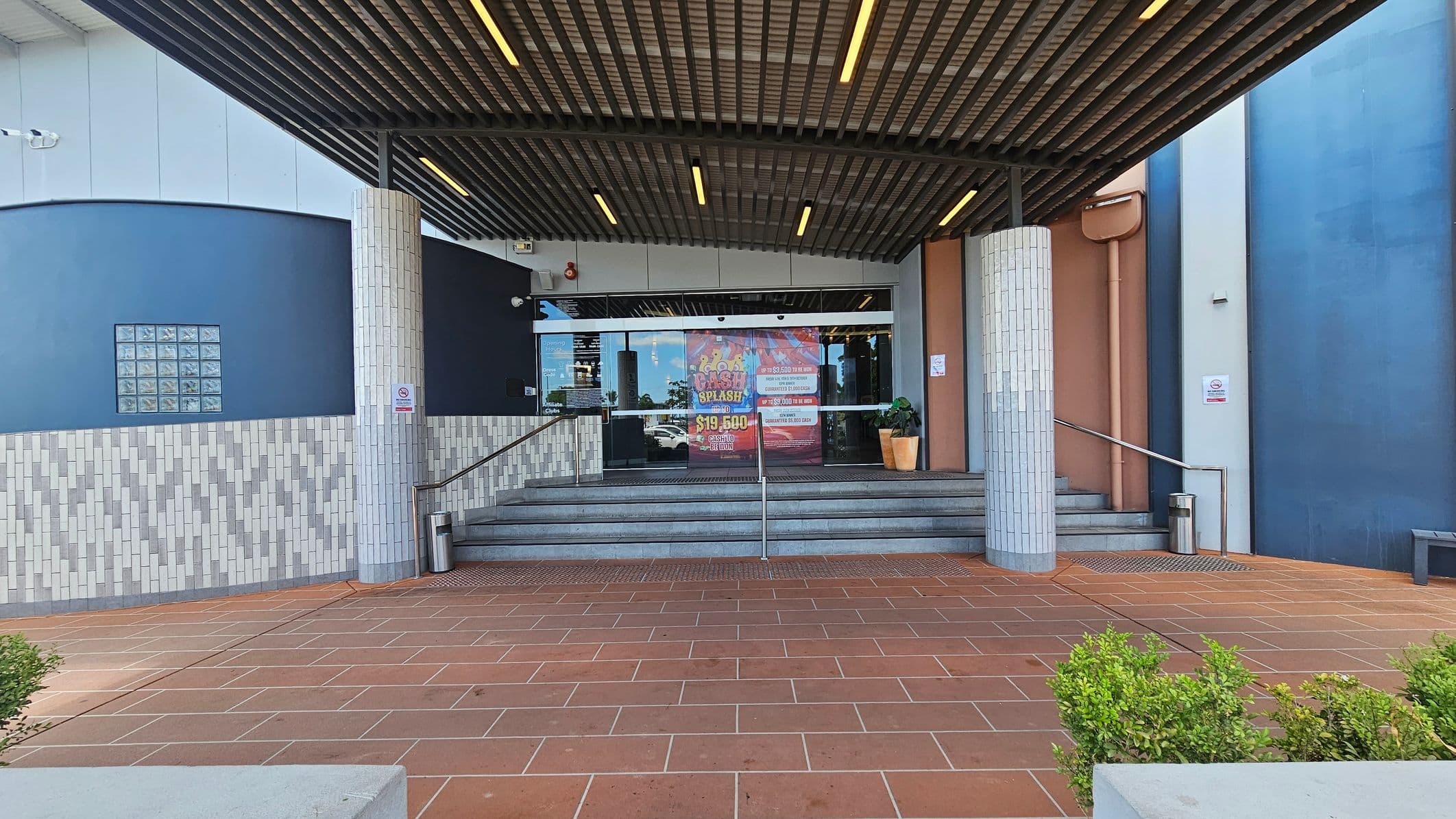 Entrance with a tiled floor leading to a set of stairs. The ceiling has a grid pattern with recessed lighting. Two cylindrical columns flank the entrance, which has glass doors displaying colourful promotional posters. The walls are a mix of blue and light grey tiles. On the right, a potted plant is placed near the door. Small bushes are visible in the foreground.