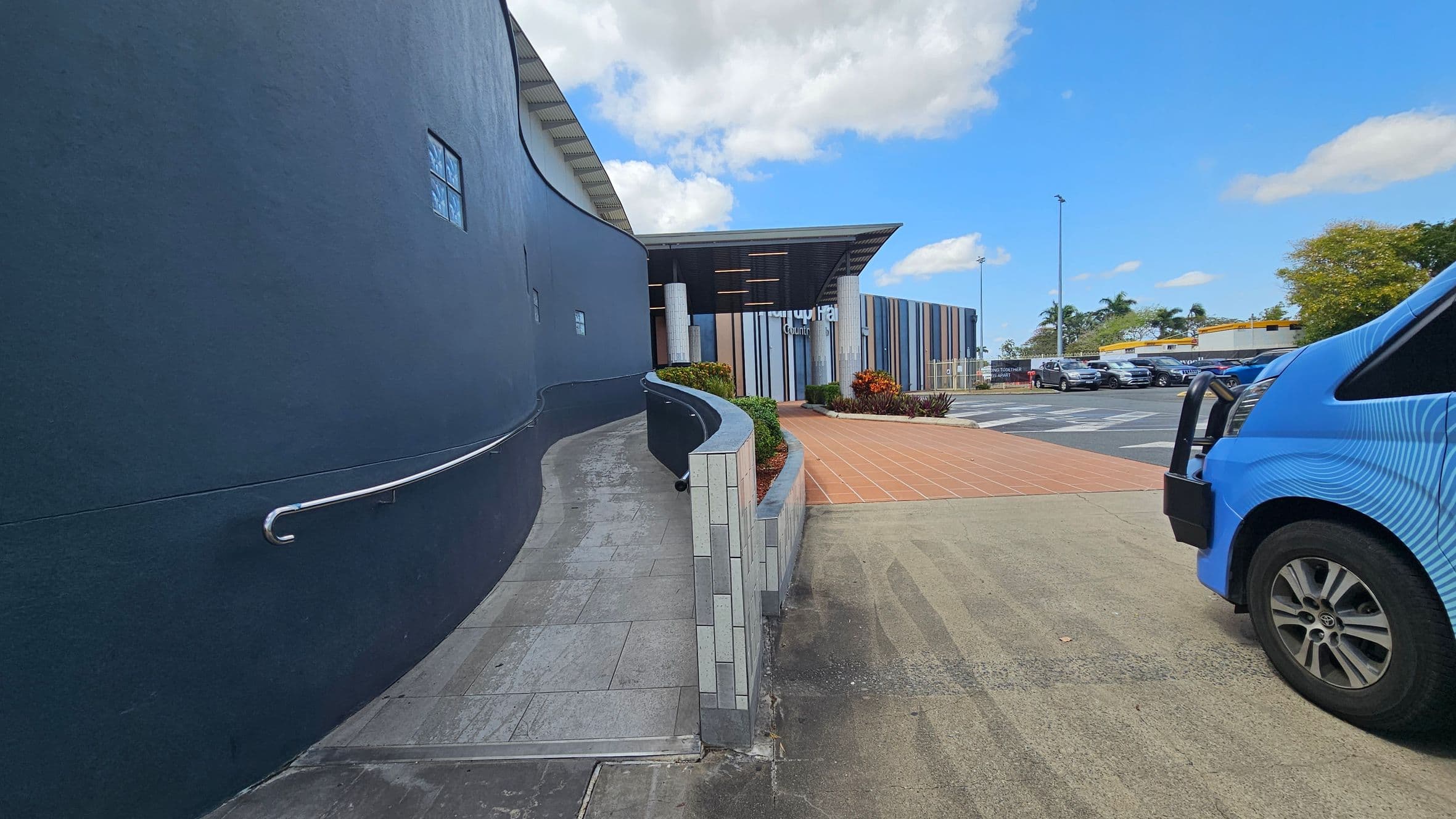 A curved, dark grey wall with a metal handrail on the left leads to a building with a striped facade in the centre. The path is tiled and bordered by a low wall with plants. On the right, a blue vehicle is partially visible, parked on a concrete surface. In the background, there are parked cars and a yellow-roofed building, with trees and a lamppost visible.