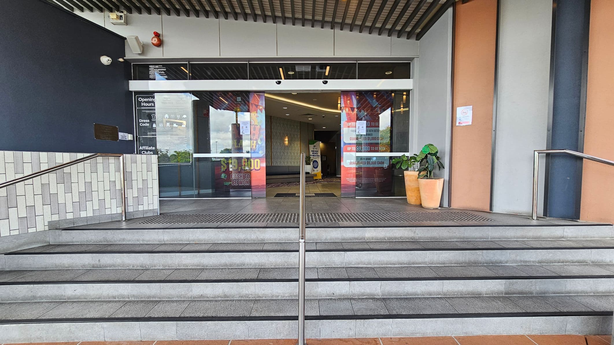 Entrance with a set of grey tiled steps leading up to glass doors. A metal handrail runs up the centre. On the left, a dark wall with a security camera and a fire alarm. Signs on the glass doors display opening hours and promotions. On the right, two potted plants sit beside the entrance. The ceiling has a slatted design, and the walls are a mix of dark and light panels.
