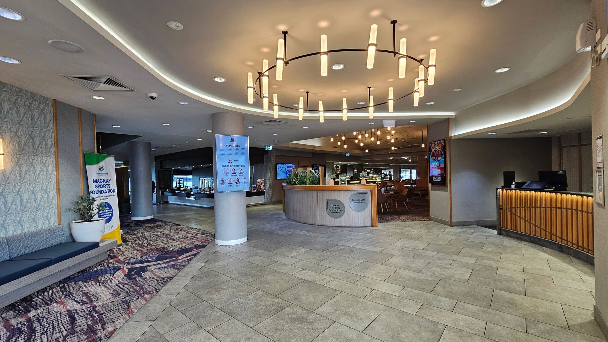 A modern interior with a tiled floor and a curved ceiling featuring recessed lighting. On the left, there's a patterned carpet, a bench, a potted plant, and a banner for the Mackay Sports Foundation. In the centre, a circular chandelier hangs above a wooden reception desk with plants. To the right, a counter with vertical wooden slats and a digital screen. In the background, tables and chairs are visible, suggesting a dining area.