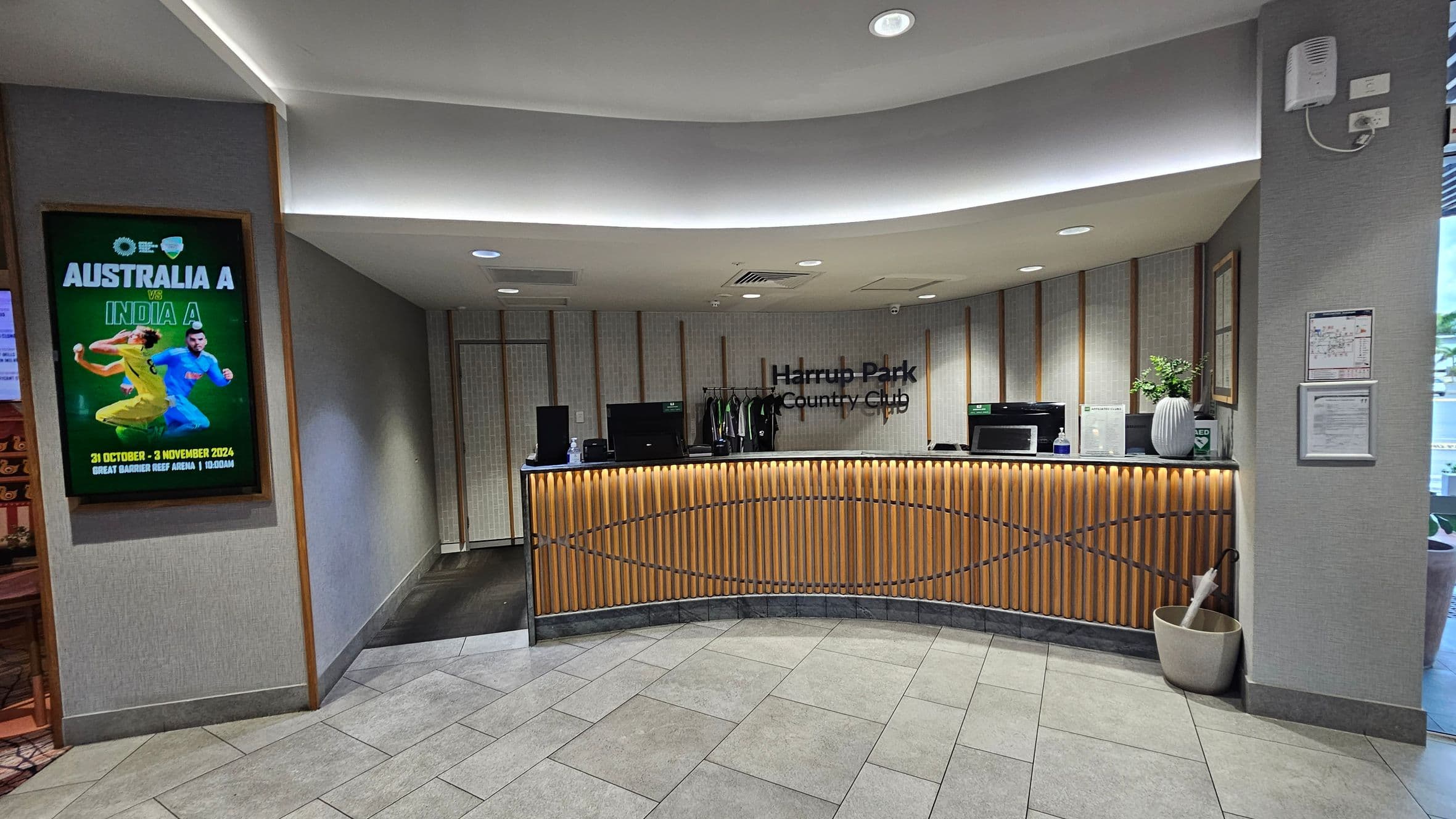 A reception area with a curved wooden desk in the centre, featuring a sign that reads "Harrup Park Country Club." Behind the desk are computers and a rack with clothing. On the left, a poster advertises a cricket match between Australia A and India A. The floor is tiled in a light grey pattern, and the ceiling has recessed lighting. On the right, a wall-mounted speaker and framed documents are visible. A potted plant and umbrella stand are near the desk.