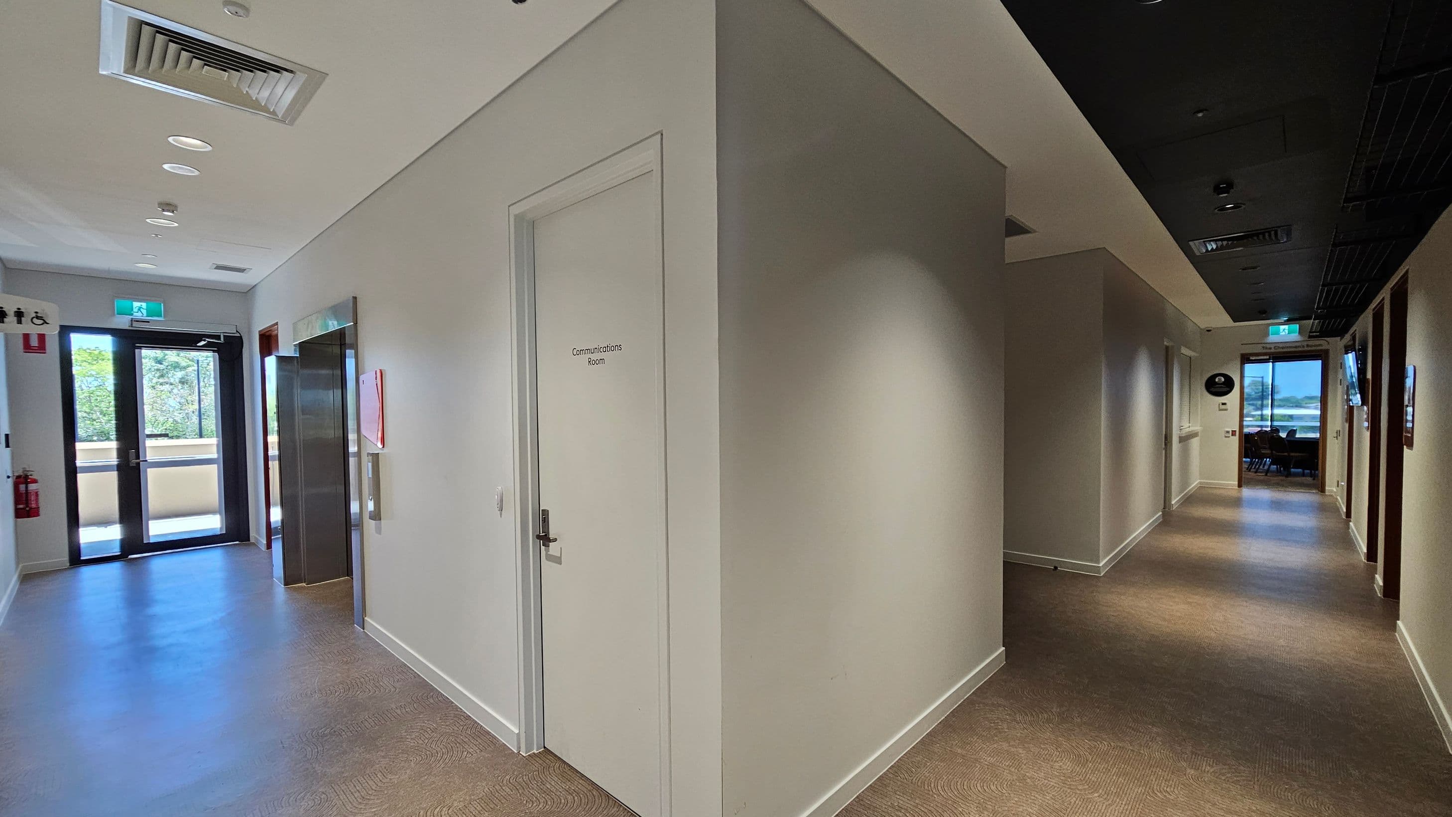 A hallway with a beige carpet and light-coloured walls. On the left, there is a door marked "Communications Room" and an elevator. Further left, a glass door leads outside, with a fire extinguisher and restroom signs nearby. On the right, a long corridor extends with several doors and a window at the end. The ceiling has recessed lighting and air vents.