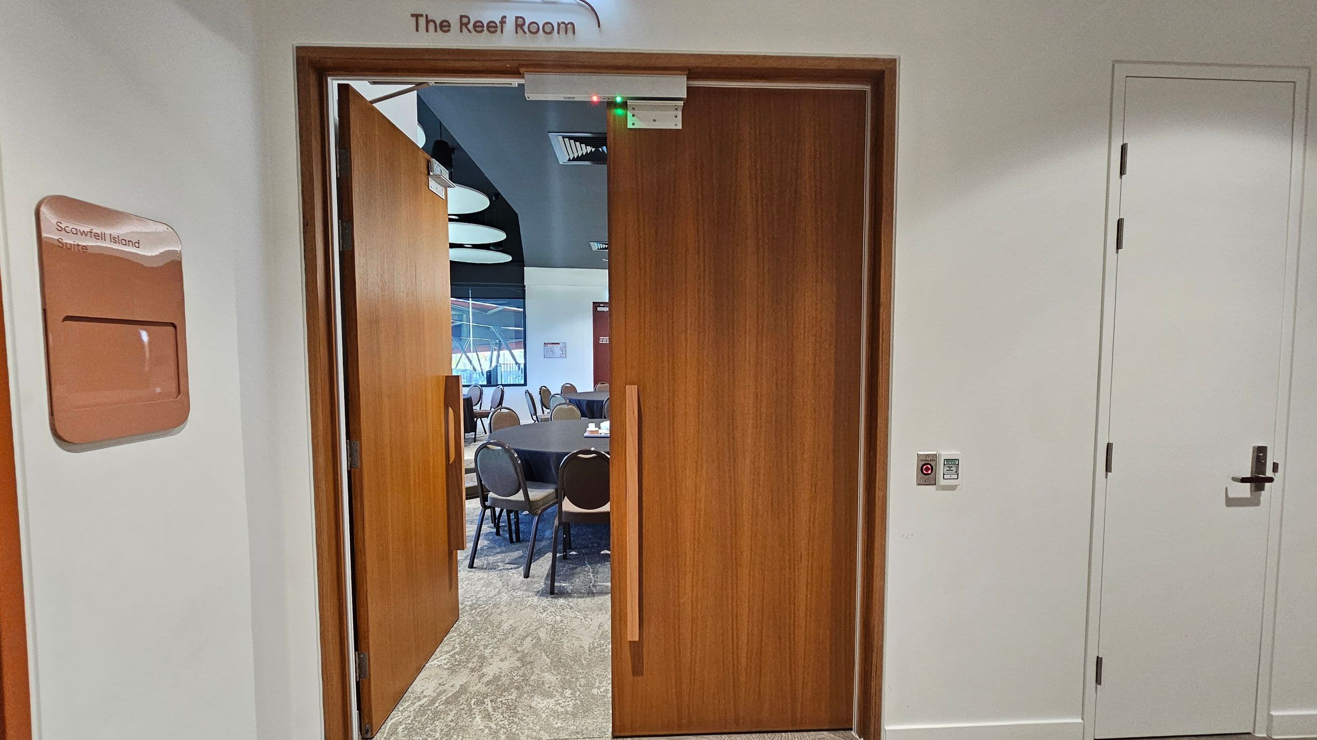 A hallway with a partially open wooden door leading into a room labelled "The Reef Room". To the left, a sign reads "Scawfell Island Suite". Inside the room, round tables and chairs are visible. The floor is carpeted, and the ceiling has circular light fixtures. On the right, a closed white door with a handle and a small control panel is mounted on the wall.