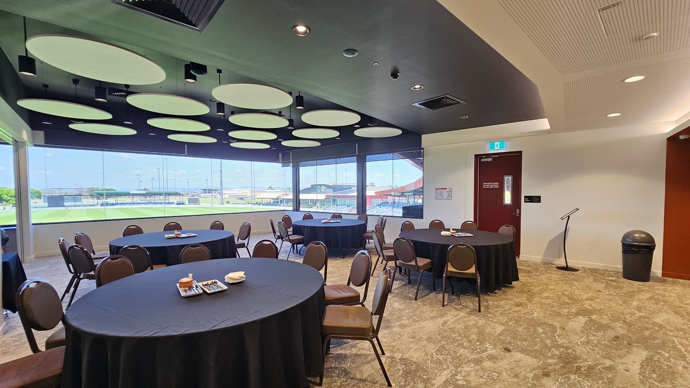 A conference room with round tables covered in black tablecloths, surrounded by brown chairs. On the tables are trays with snacks and napkins. The left side features large windows with a view of a sports field. The ceiling has circular light fixtures. On the right, there's a red door, a microphone stand, and a bin. The floor is carpeted in a grey pattern.