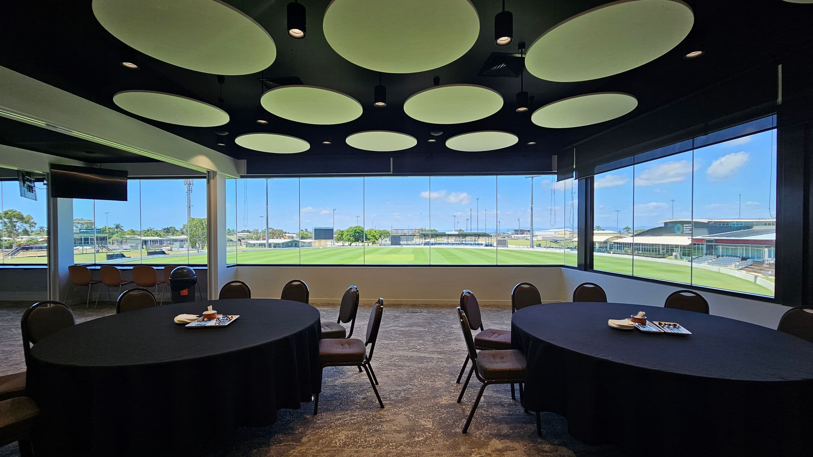 A room with large windows overlooking a sports field. In the foreground, there are round tables covered with black tablecloths and surrounded by chairs. On the tables are trays with snacks. The ceiling features circular light fixtures. In the background, the view includes a green field, a scoreboard, and buildings. The floor is carpeted with a patterned design.