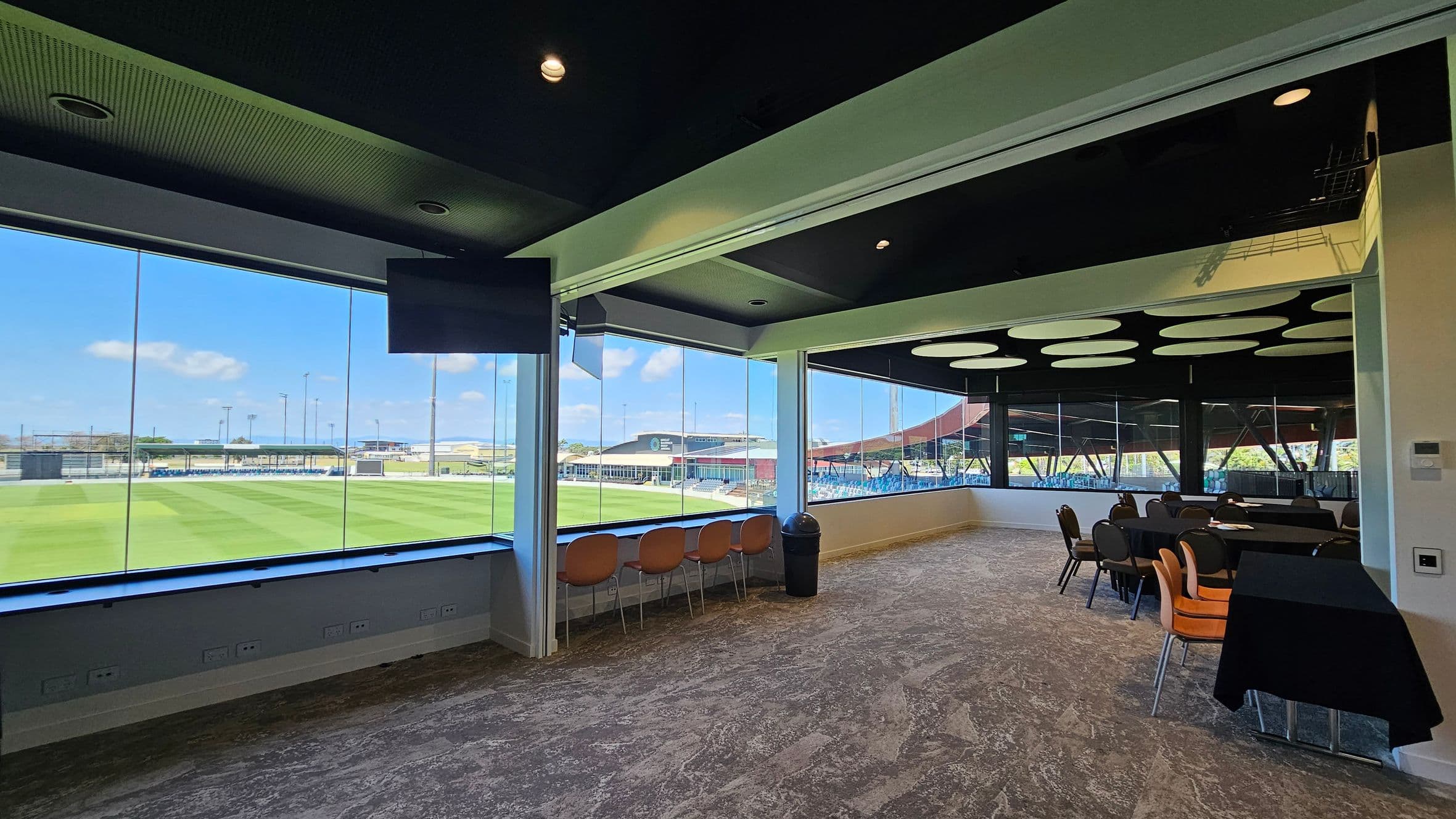 A room with large glass windows offering a view of a sports field. On the left, there are orange chairs lined up against the window. On the right, there are tables covered with black cloths and surrounded by chairs. The ceiling is dark with circular light fixtures, and the floor has a patterned carpet. A black bin is placed near the windows.