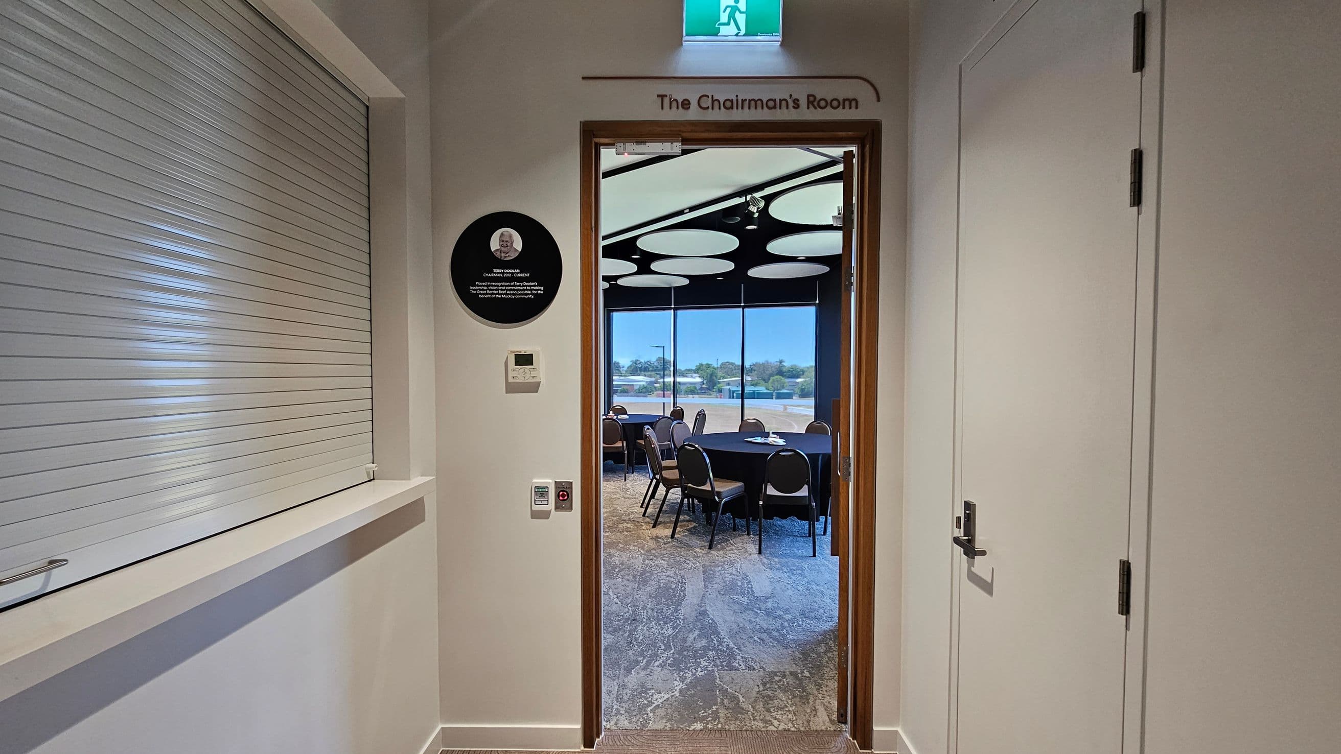 A hallway with a door labelled "The Chairman's Room" in the centre. To the left, there's a closed shutter window and a wall plaque. On the right, a closed white door with a handle. Through the open door, a room with round tables and chairs is visible, with large windows showing an outdoor view. The ceiling inside the room has circular light fixtures. The floor is carpeted with a patterned design. An exit sign is above the door.
