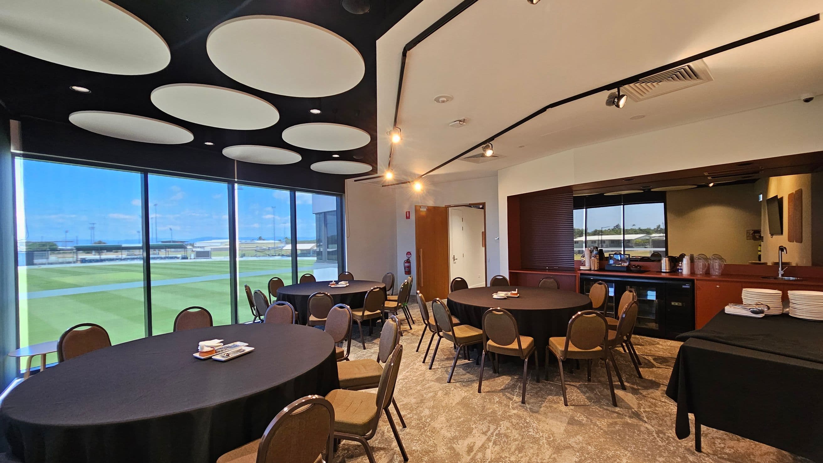 A conference room with round tables covered in black tablecloths, surrounded by brown chairs. On the left, large windows offer a view of a green field. The ceiling features circular light fixtures. On the right, a counter with plates, cups, and a coffee machine is visible. The floor is carpeted with a patterned design, and there is a fire extinguisher near a wooden door.