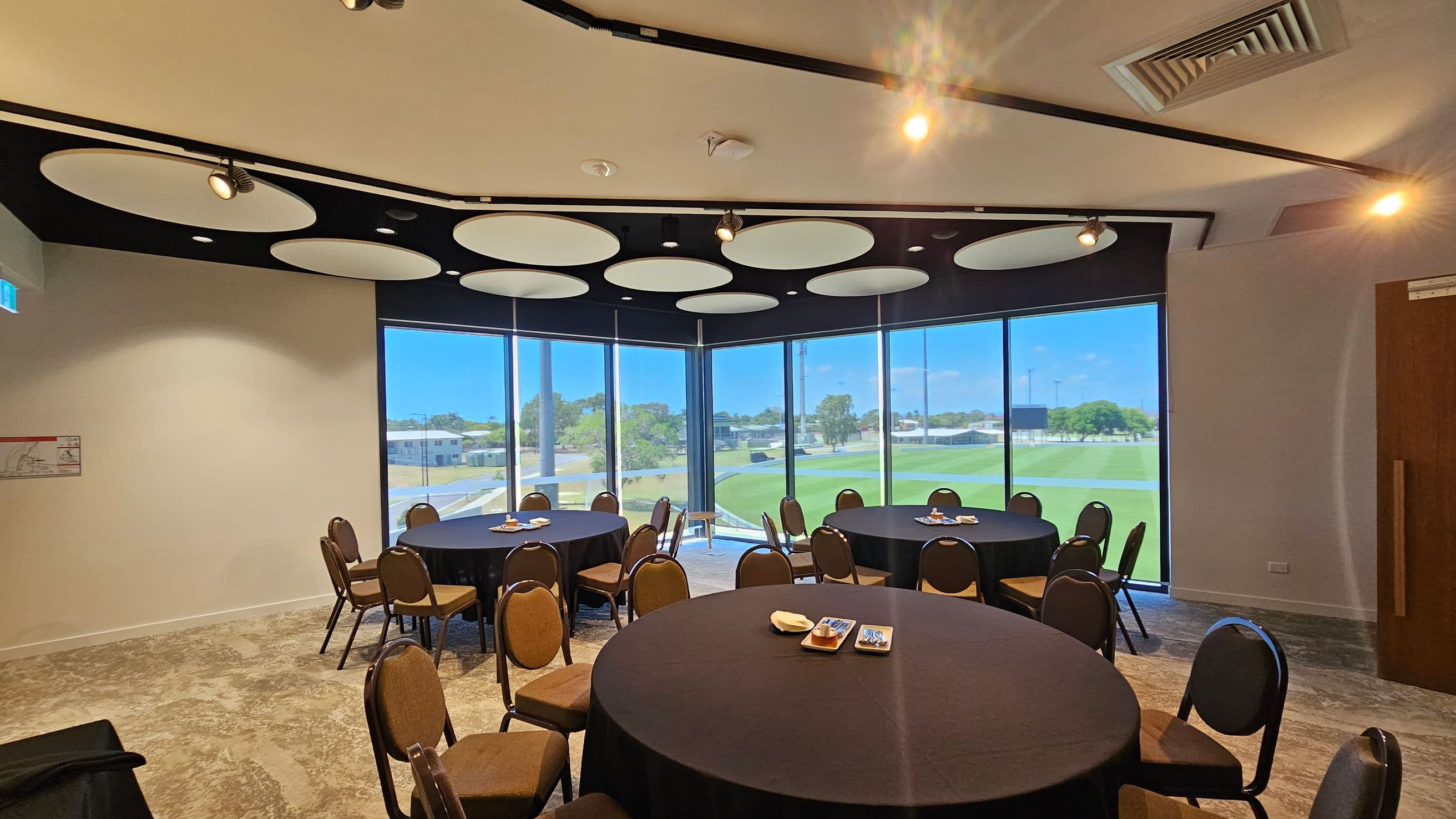 A conference room with round tables covered in black tablecloths, surrounded by brown chairs. Each table has small plates with snacks. Large windows on the right and centre offer a view of a green outdoor area with trees and buildings. The ceiling features circular light fixtures and air vents. The floor is carpeted in a neutral tone. A door is visible on the right, and an emergency exit sign is on the left wall.