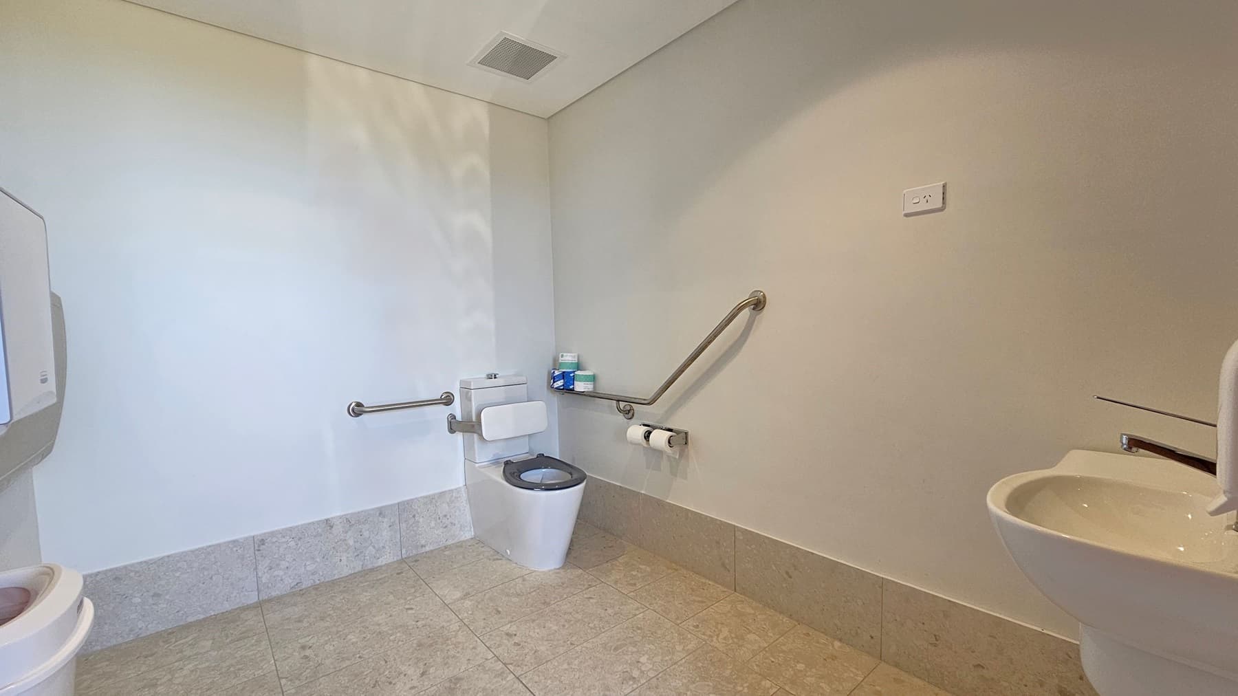 A spacious, accessible bathroom with a white toilet in the centre, equipped with stainless steel grab bars on both sides. To the right, a white sink is partially visible. On the left, a wall-mounted paper towel dispenser is present. The walls are light-coloured, and the floor is tiled in a light grey pattern. A power outlet is visible on the right wall. The ceiling is plain with a vent.