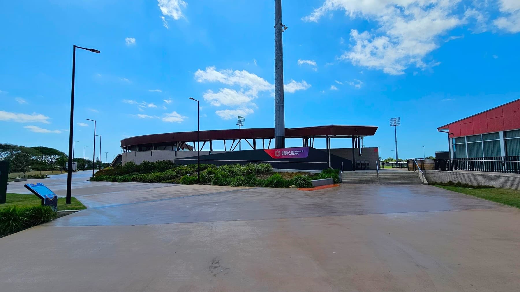A wide concrete pathway leads to the Great Barrier Reef Arena, with a large sign in the centre. The arena has a modern design with a circular roof and visible floodlights. On the left, there are street lamps and a grassy area with trees. On the right, a red building with large windows is visible. The area is landscaped with shrubs and plants near the entrance.
