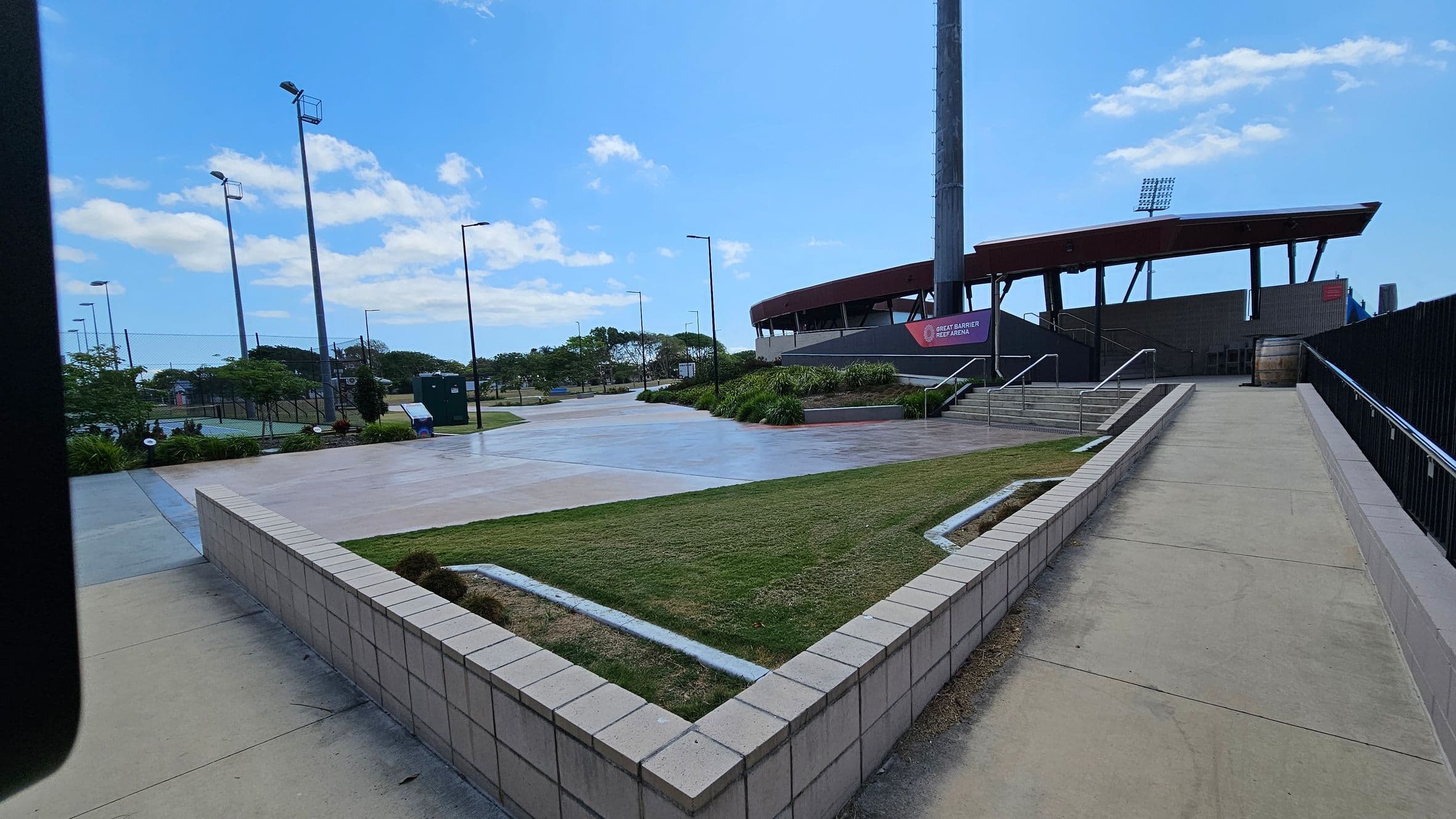 A paved walkway leads up to a stadium on the right, with a sign reading "Great Barrier Reef Arena." The stadium has a modern design with a slanted roof and large floodlights. On the left, there are several tall light poles and a landscaped area with grass and small shrubs. The foreground features a low brick wall bordering a grassy patch. The background includes more greenery and a clear view of the stadium structure.