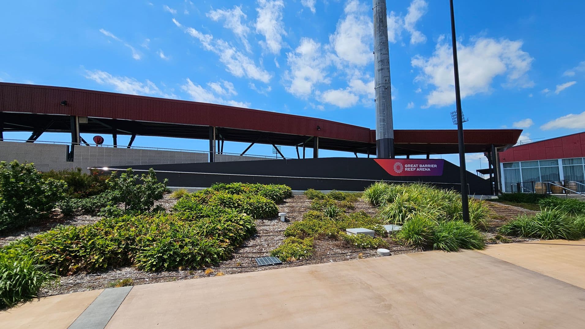 A sports arena with a red and grey exterior. In the foreground, there are landscaped gardens with various green shrubs and plants. A tall light pole stands near a sign that reads "Great Barrier Reef Arena." The ground is paved with a light brown surface. The arena structure features open sections supported by black beams.