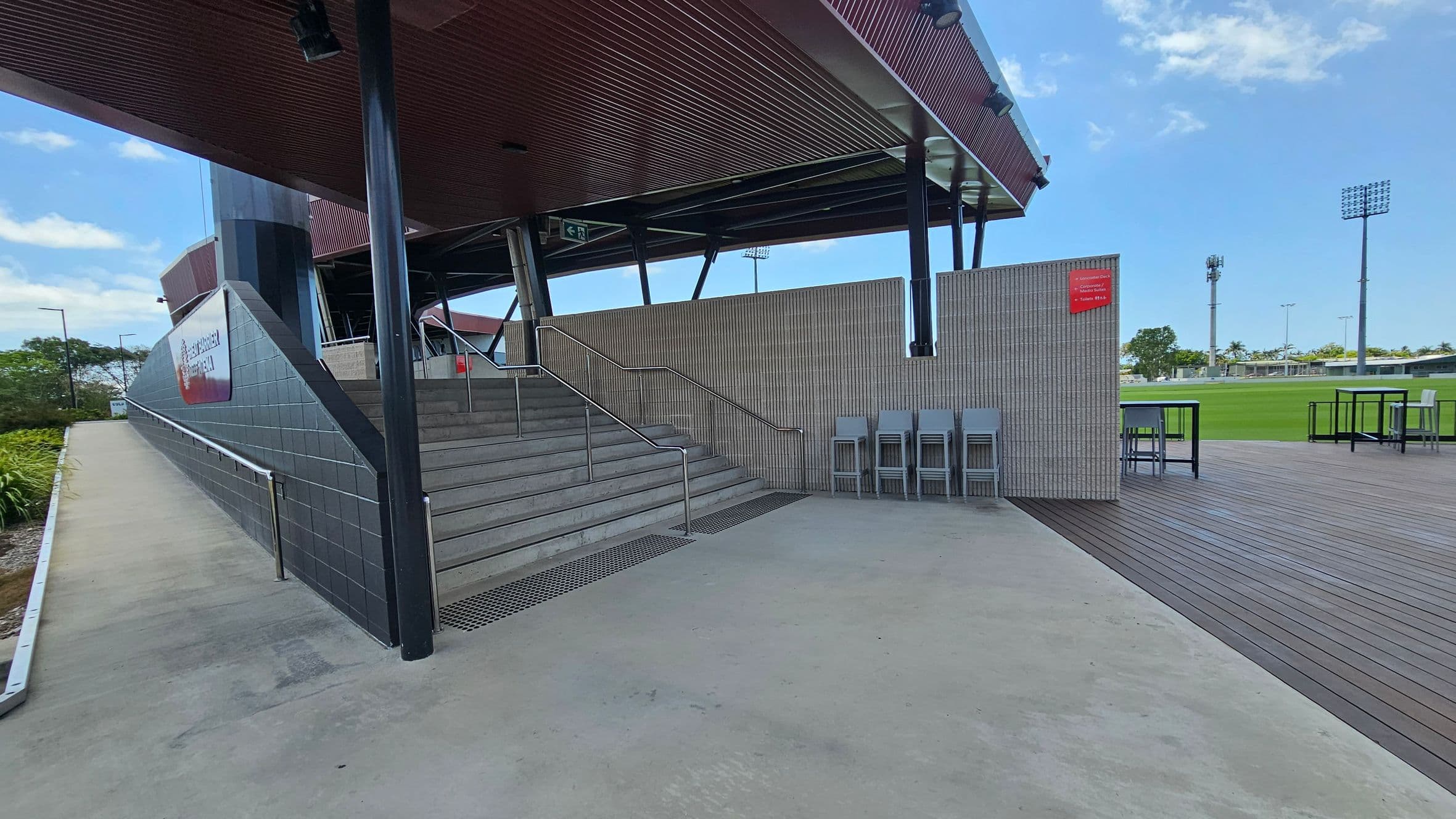Concrete steps with metal railings lead up to a covered area supported by dark pillars. To the right, a wooden deck features several high tables and chairs. A grey tiled wall with a red sign is adjacent to the steps. In the background, a green field is visible with tall floodlights and trees. The ceiling is dark and slatted, and the floor is a mix of concrete and wood.