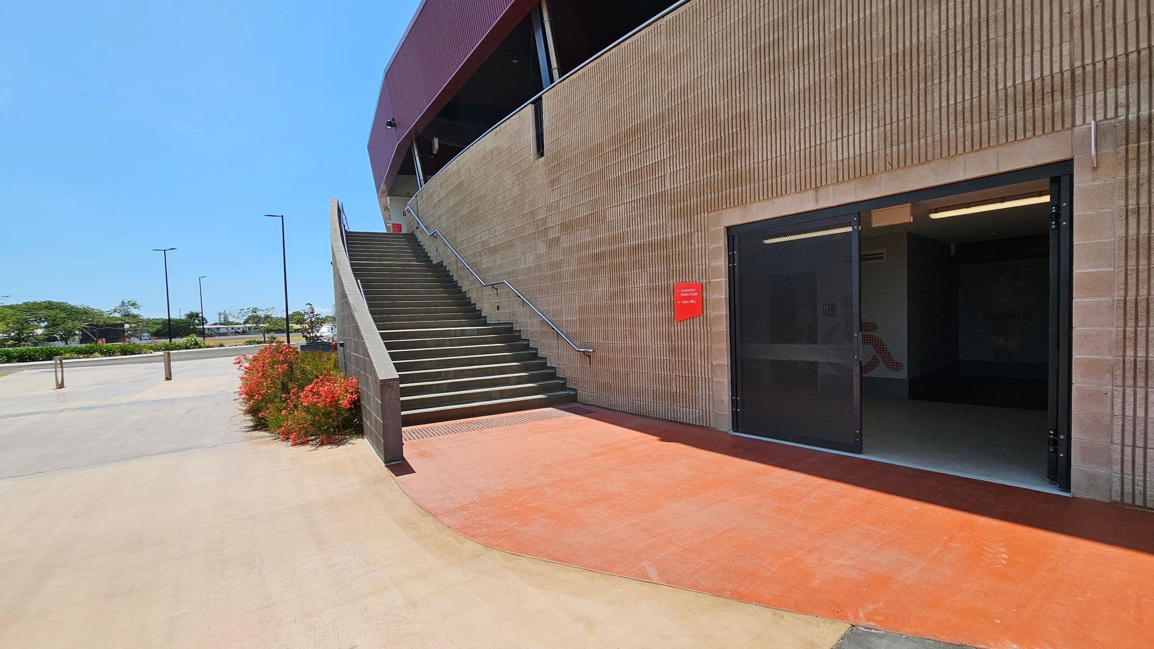 On the right, there is a building with a textured brick wall and a large open doorway. A red sign is mounted on the wall. A staircase with a metal railing ascends alongside the building. On the left, there is a paved area with red flowering bushes and several streetlights. The background features trees and a distant view of a road. The ground is a mix of red and beige concrete.