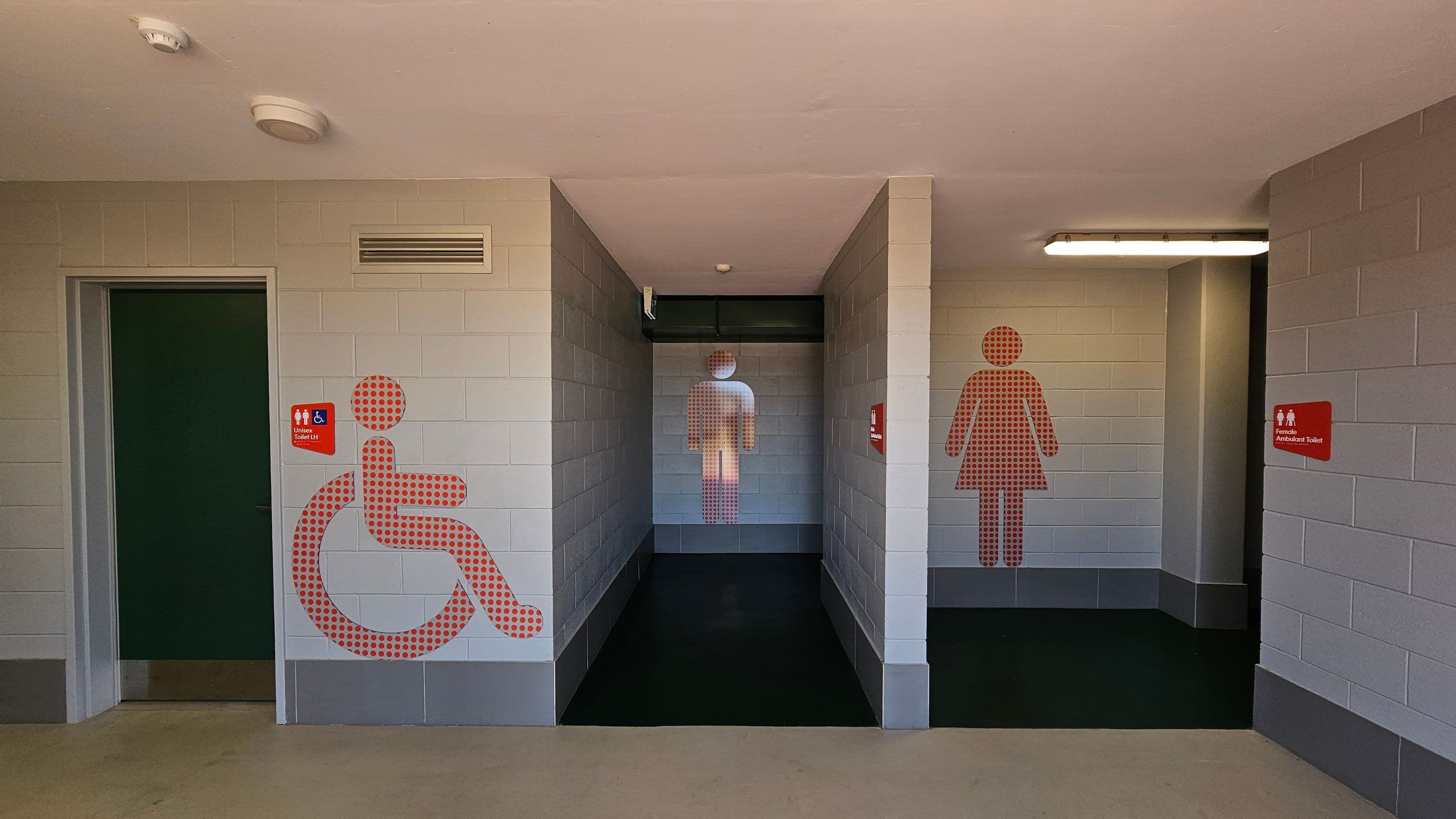 A public toilet entrance with three sections. On the left, a door with a large red dotted wheelchair symbol and a sign indicating a unisex toilet. In the centre, a corridor with a red dotted male figure on the wall. On the right, a red dotted female figure next to a sign for a female ambulant toilet. The walls are light grey with darker grey at the bottom, and the floor is a mix of beige and black. The ceiling is plain with two light fixtures.