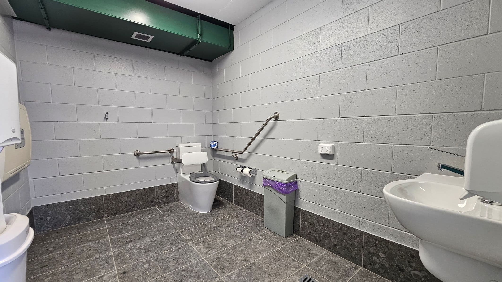 A public restroom with grey brick walls and a dark tiled floor. On the left, there's a paper towel dispenser. In the centre, a toilet with grab bars on both sides and a toilet roll holder. To the right, a bin with a purple liner and a wall-mounted sink with a soap dispenser. Above, a green ventilation duct is visible on the ceiling.