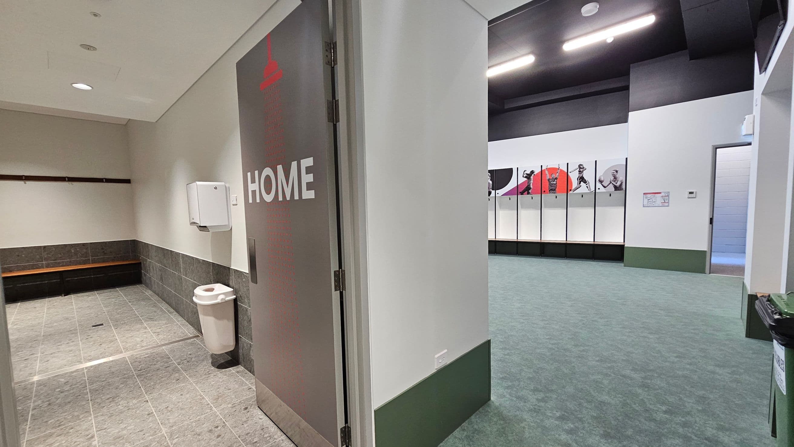 A sports locker room with a door labelled "HOME" on the right. To the left, there's a tiled area with a bench and a wall-mounted dispenser above a bin. The floor is tiled in this section. In the background, the main room has a green carpet and features lockers with sports-themed graphics. The ceiling is dark with bright lights.
