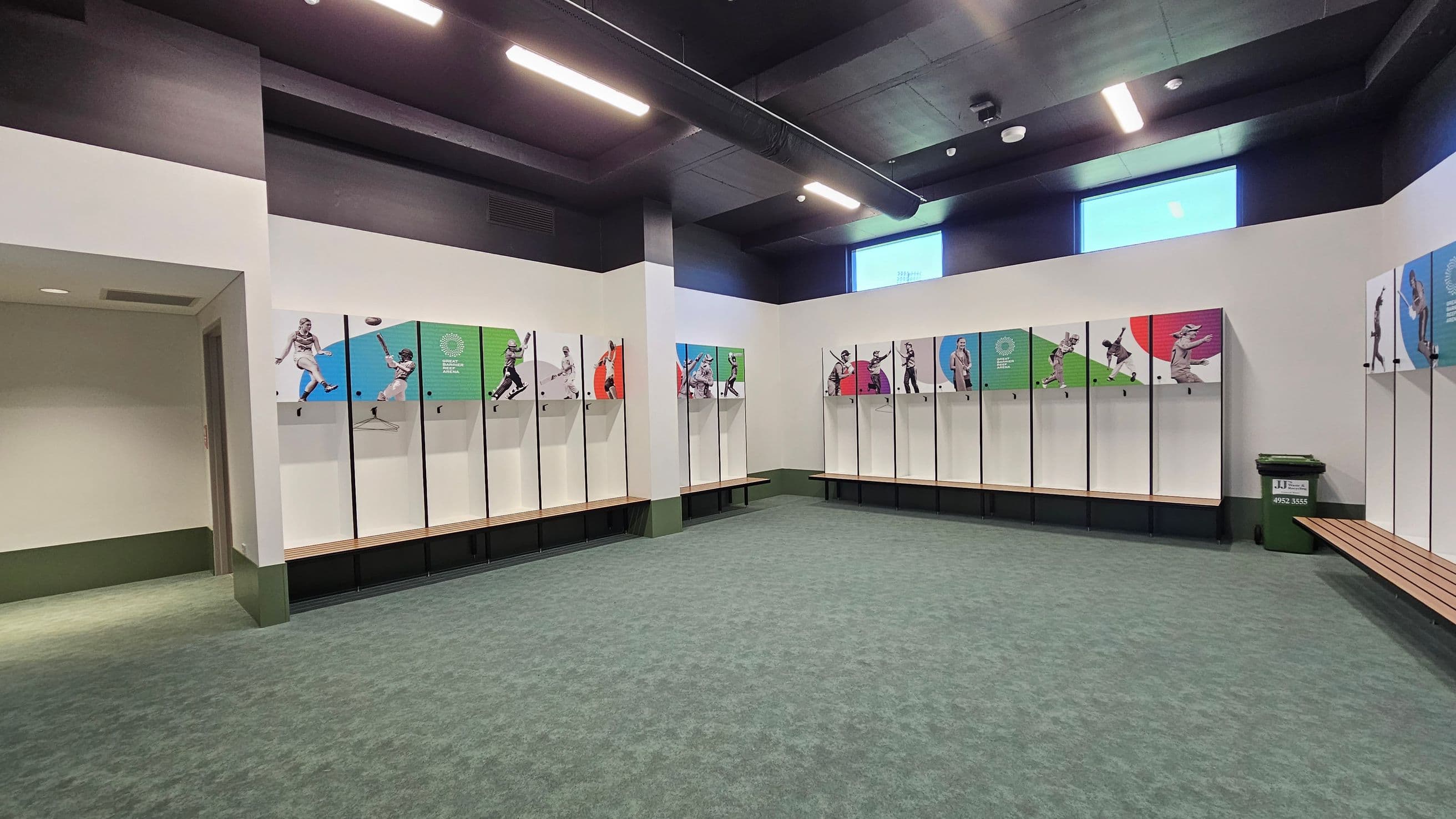 A spacious locker room with a grey and green patterned carpet. On the left, there are lockers with images of athletes in action. The back wall also features similar lockers with colourful panels. A green recycling bin is positioned on the right. The ceiling is dark with bright overhead lights, and there are two rectangular windows high on the back wall. A wooden bench is visible on the right side.