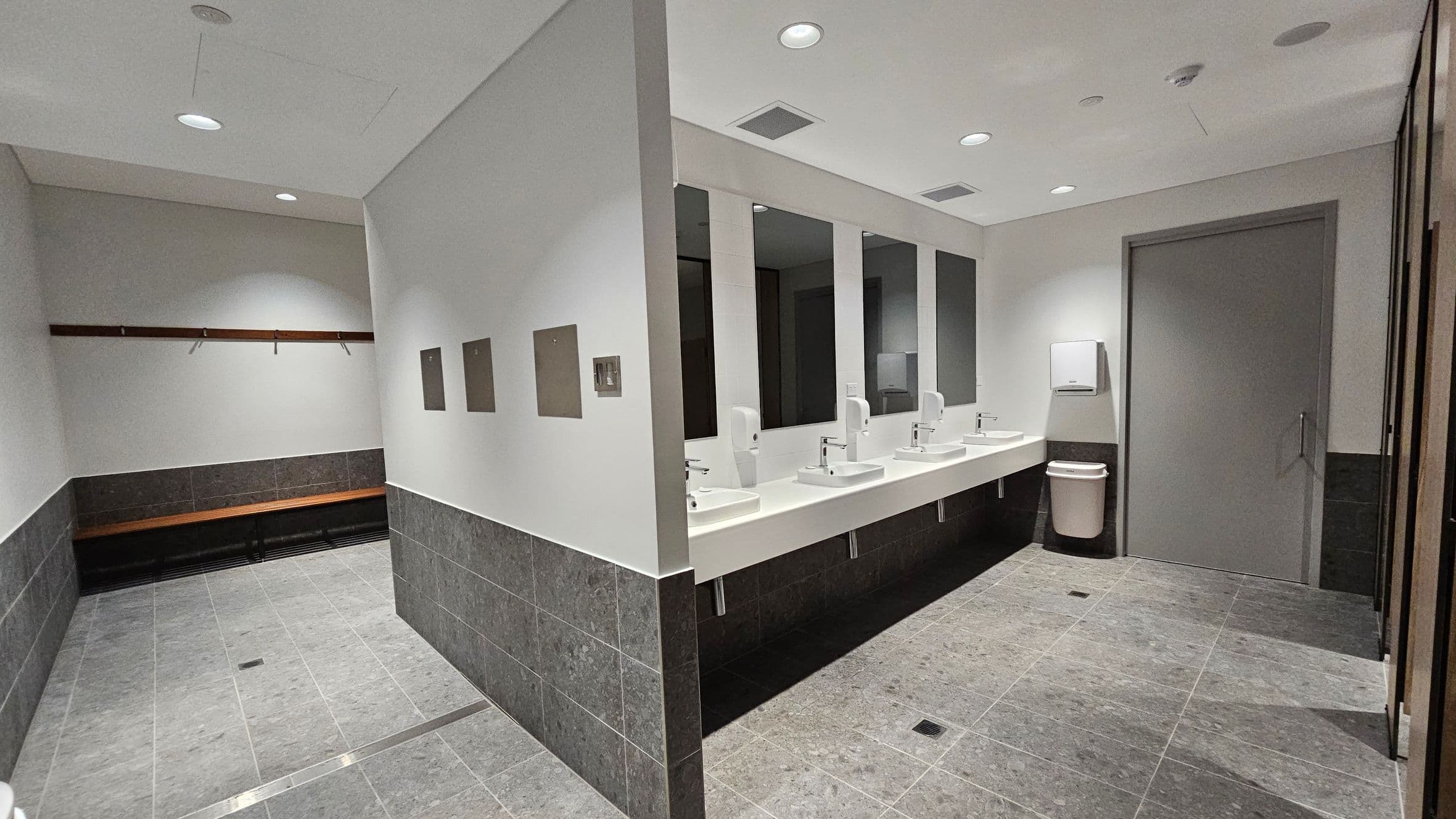 A modern public restroom with grey tiled flooring and walls. On the right, there are four sinks with mirrors above them, each equipped with a soap dispenser. A paper towel dispenser and a bin are near a closed door. On the left, a wooden bench runs along the wall with coat hooks above it. The ceiling is white with recessed lighting.