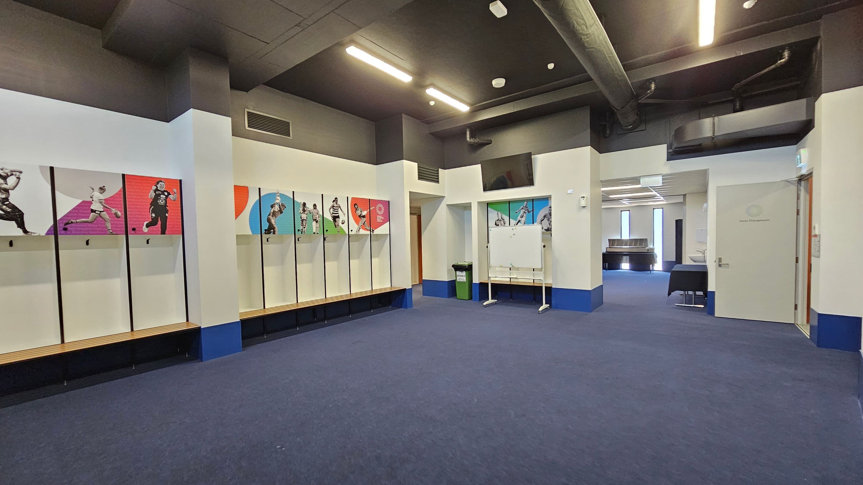 A spacious locker room with a blue carpeted floor. On the left, there are lockers with colourful panels featuring sports figures. In the centre, a whiteboard and a green bin are visible. The ceiling has exposed ducts and bright lights. On the right, an open door leads to another room with a table and chairs. A wall-mounted TV is above the doorway.