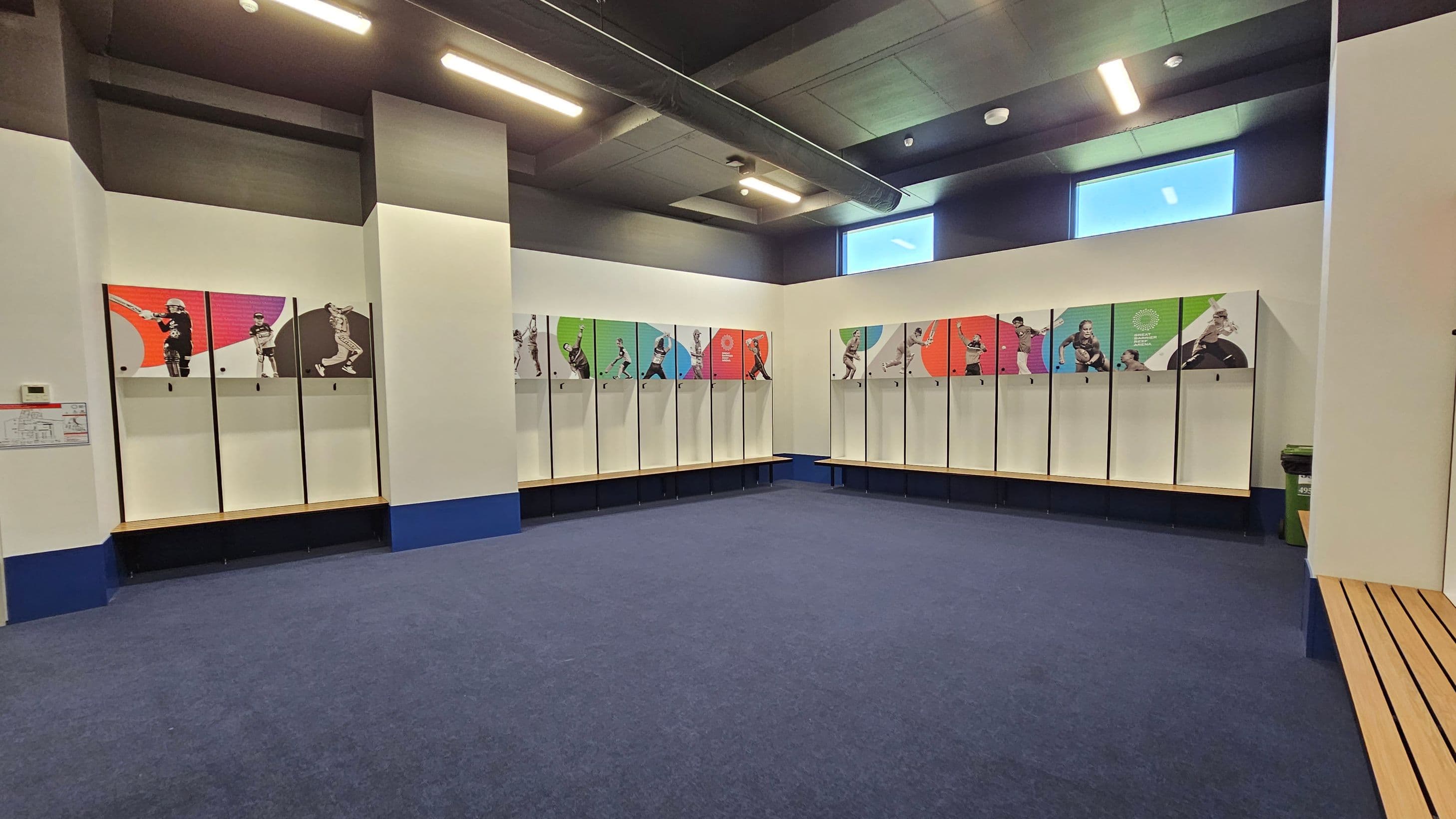 A spacious locker room with a blue carpeted floor. Lockers line the walls, each featuring colourful graphics of athletes in action. The lockers have a white base with a wooden bench below. The ceiling is dark with exposed ductwork and bright overhead lights. Two small windows near the ceiling allow natural light in. A recycling bin is visible on the right side.