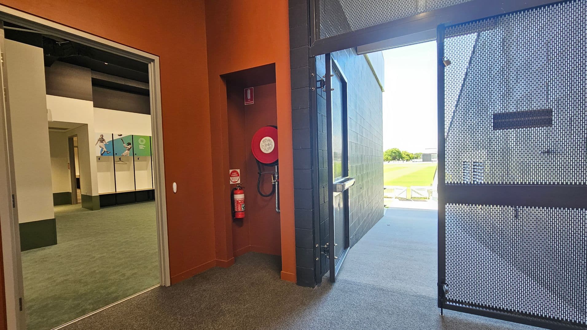 On the left, there's an open doorway leading to an interior space with green carpet and sports-themed wall panels. In the middle, a fire hose reel and extinguisher are mounted on a reddish-brown wall. On the right, a metal mesh door is open, leading outside to a grassy area with a clear view of the outdoors. The floor is carpeted inside and transitions to a concrete surface outside.