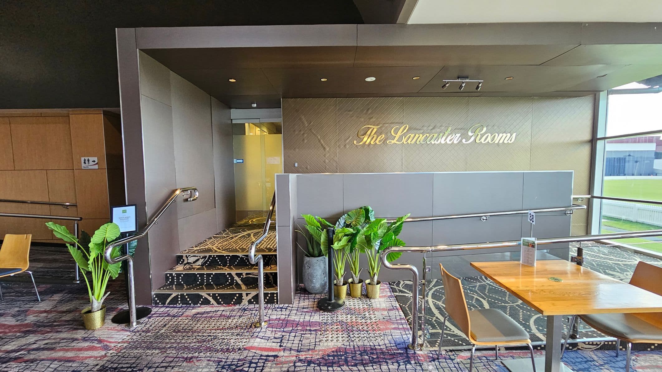 Entrance to "The Lancaster Rooms" with a sign on the wall. On the left, a staircase with a metal railing leads up, flanked by potted plants. A sign indicates restrooms. The floor is covered with a patterned carpet. On the right, a wooden table with chairs is positioned near a large window, offering a view of a green outdoor area. The ceiling is dark with recessed lighting.