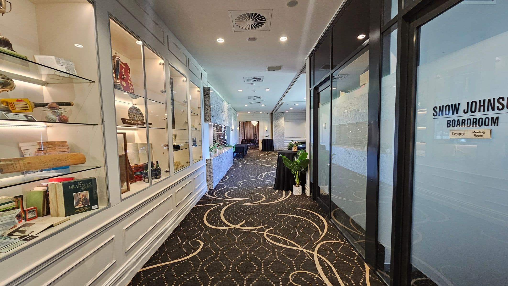 On the left, there is a glass display case filled with various memorabilia, including books, a cricket bat, and trophies. The right side features a glass wall with a sign reading "Snow Johnson Boardroom" and an "Occupied" notice. The floor is carpeted with a geometric and swirling pattern, and the ceiling has recessed lighting and air vents. In the background, there are tables with black tablecloths and a potted plant.