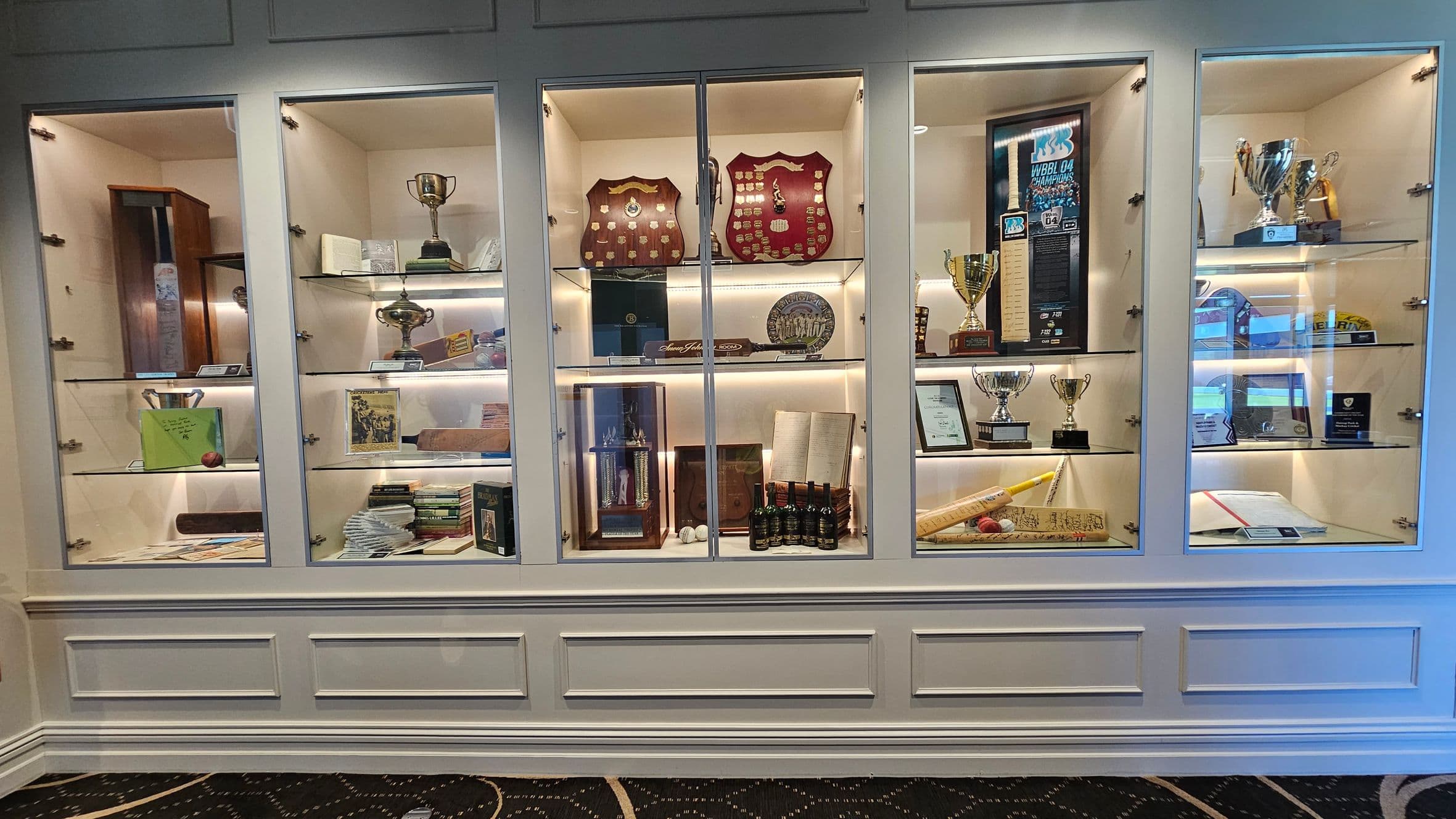 A display cabinet with multiple glass sections showcasing various trophies, plaques, and memorabilia. On the left, there are trophies and books. The centre features shields, a trophy, and bottles. On the right, more trophies and a cricket bat with balls are visible. The cabinet is set against a white wall with a patterned carpeted floor.