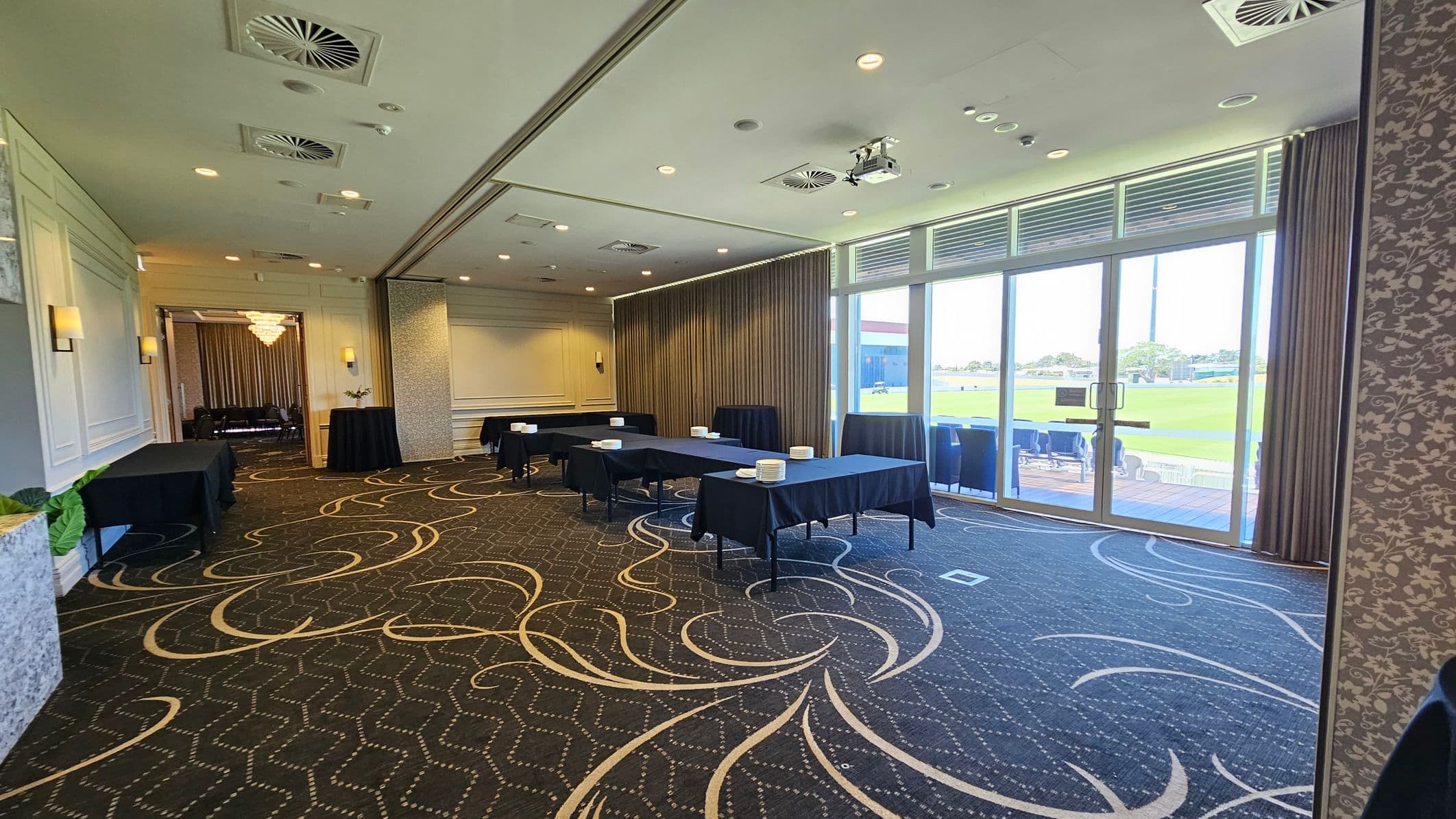 A conference room with patterned carpet and a ceiling with recessed lighting and air vents. On the left, there are black-draped tables and a doorway leading to another room with a chandelier. In the centre, several tables are arranged with plates and napkins. On the right, large glass doors with curtains open to a view of a green outdoor area. The walls are decorated with light-coloured panels and floral wallpaper.