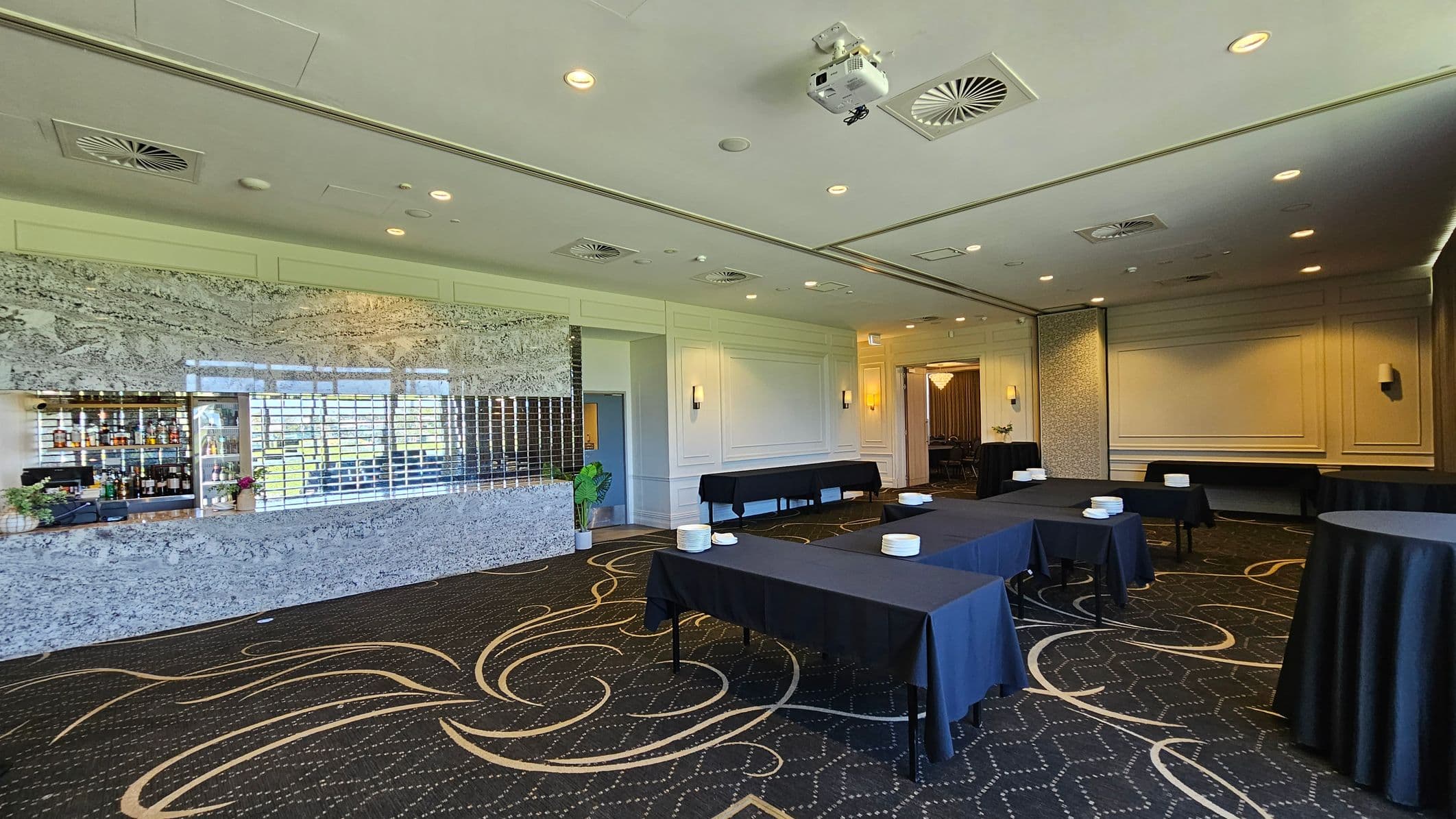 A spacious room with a patterned dark carpet featuring swirling designs. On the left, there's a bar area with a mirrored backdrop and shelves stocked with bottles. In the centre, several tables are covered with black tablecloths, each with stacks of white plates. The walls are decorated with moulding and sconces. The ceiling is equipped with recessed lighting, air vents, and a projector. A doorway on the right leads to another room.