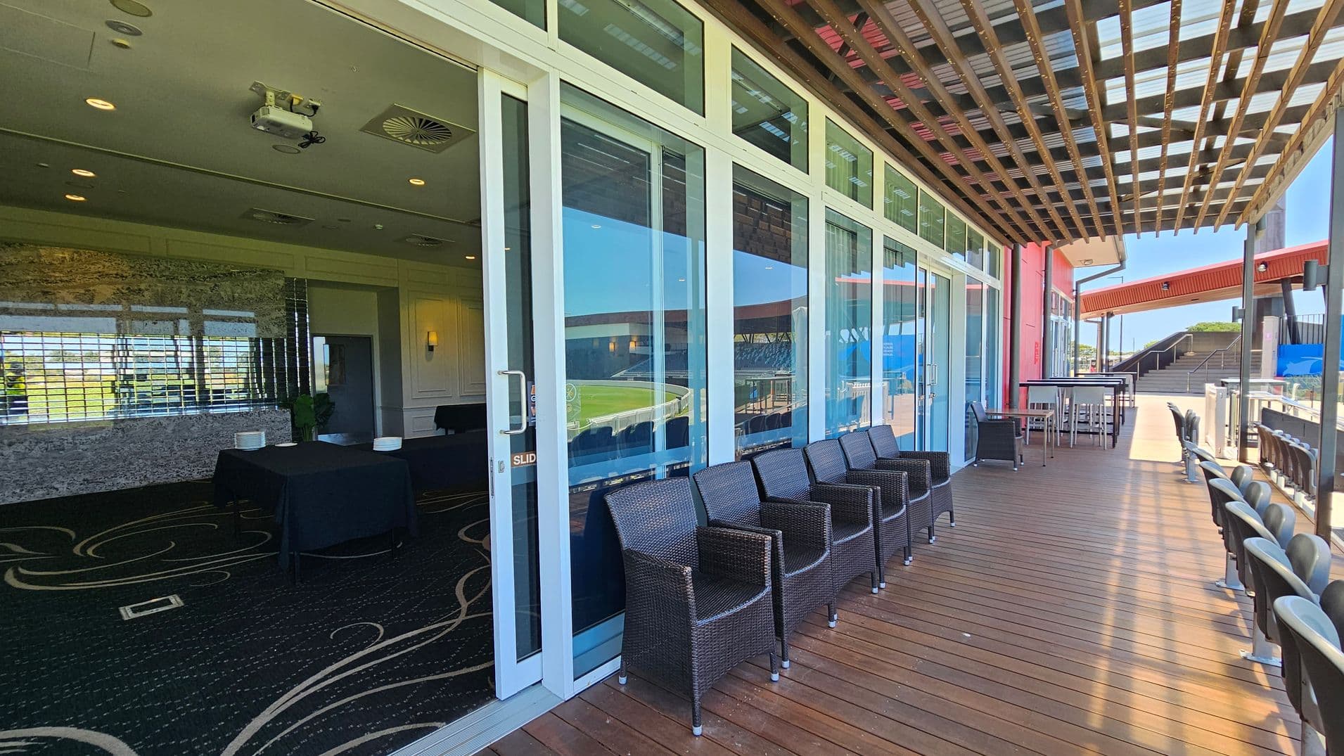 A wooden deck with wicker chairs lined up against large glass sliding doors on the left. Inside, a room with patterned carpet, tables, and a mirrored wall is visible. On the right, the deck extends with more seating and a high table with stools. Above, a wooden pergola provides partial shade. In the background, a red structure and stairs are visible, leading to an open area.