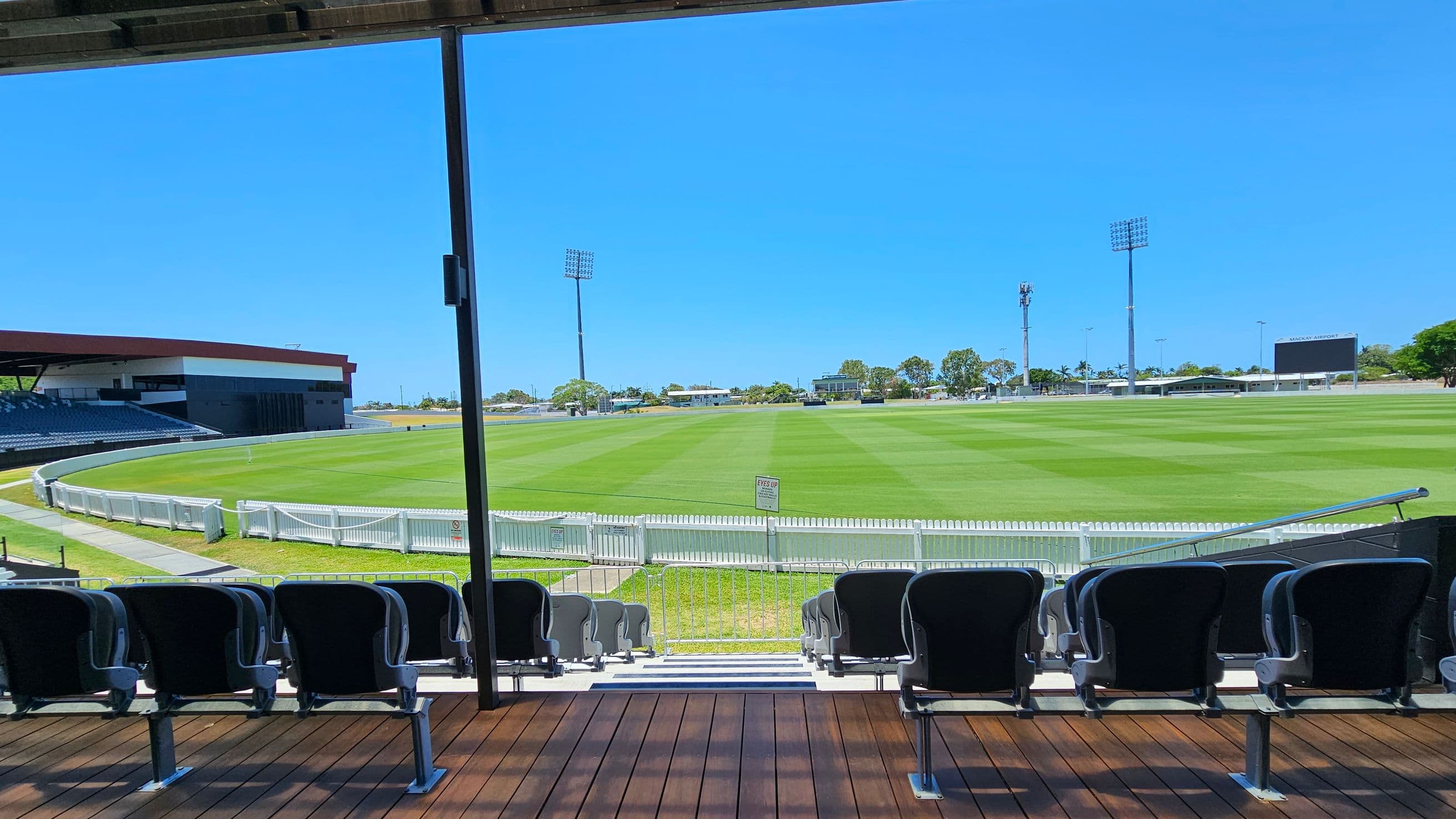 Rows of black stadium seats on a wooden platform in the foreground, facing a large, well-maintained green sports field. A white picket fence surrounds the field. In the background, there are tall floodlights and a scoreboard on the right. A large building with a red roof is on the left, and trees and smaller structures are visible in the distance. The ceiling is partially visible, with metal beams overhead.